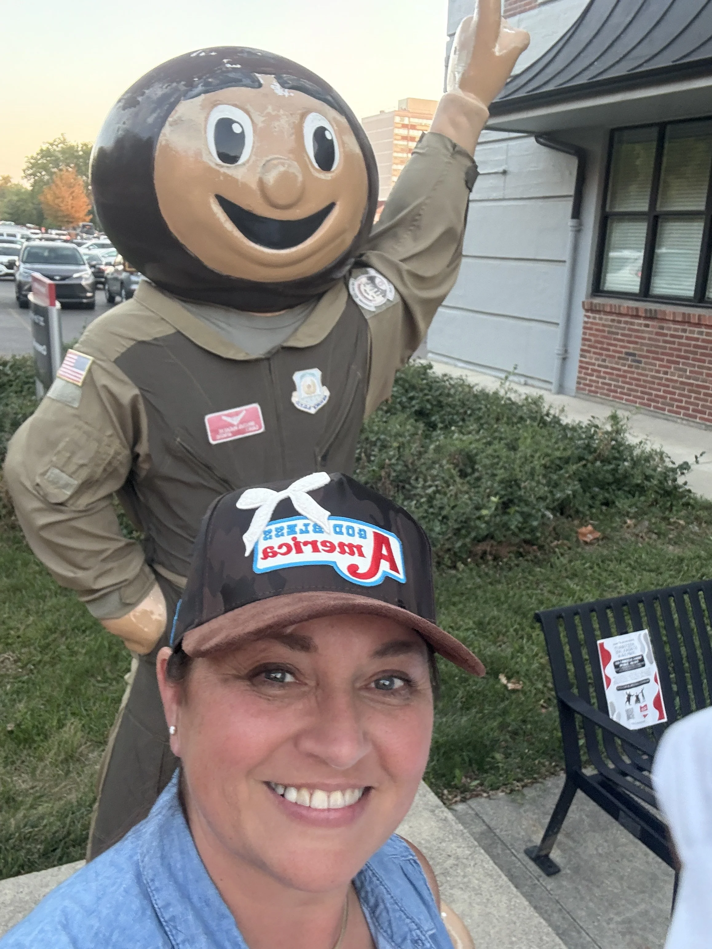 A woman smiling in the foreground with a person in a mascot costume behind her. The mascot has a large round head with a smiling face and wears a brown uniform with patches. The scene is outdoors near a parking lot and a brick building.