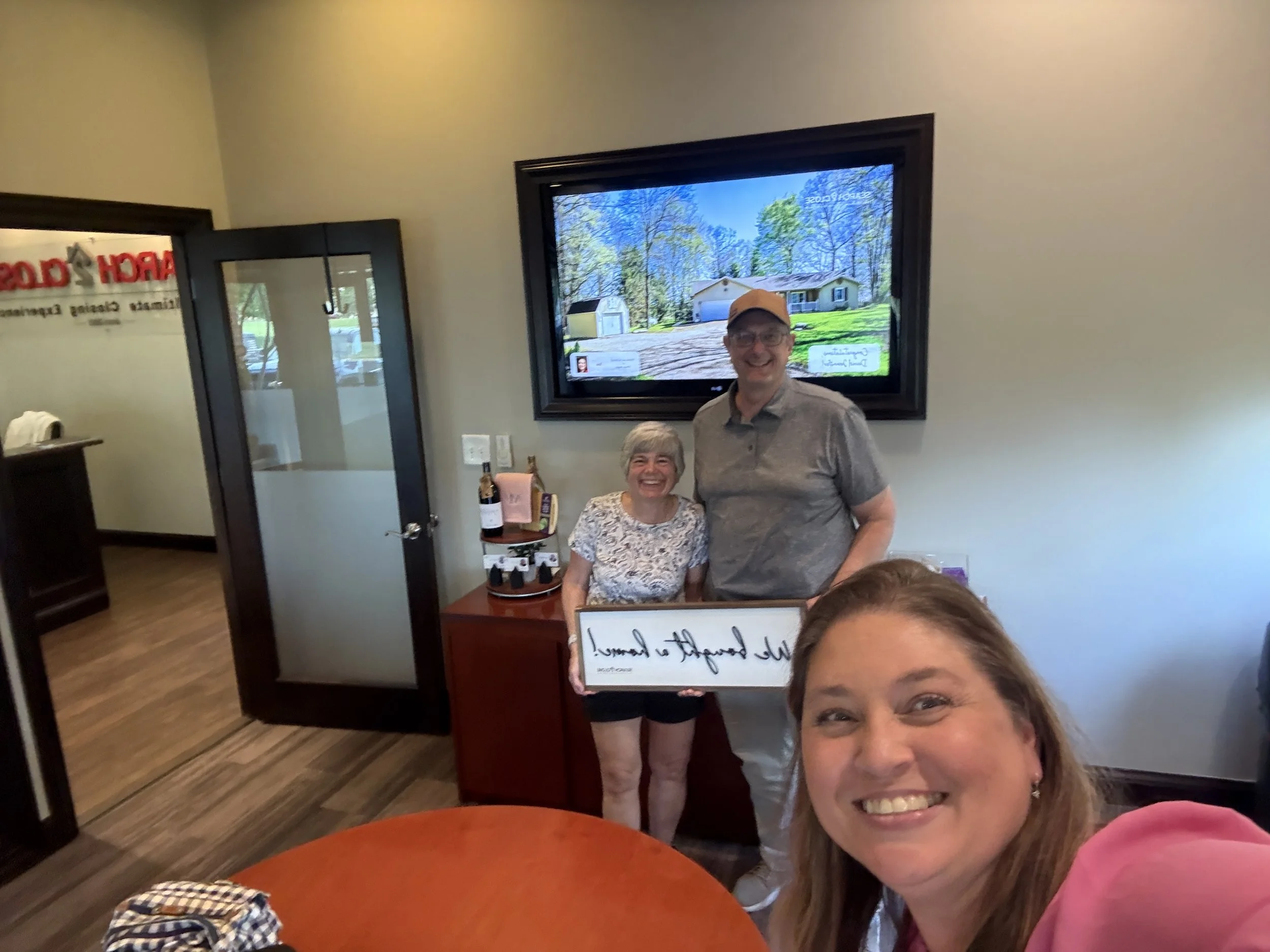 Two women smiling, one holding a framed sign, in a room with a large TV screen displaying a house and trees.
