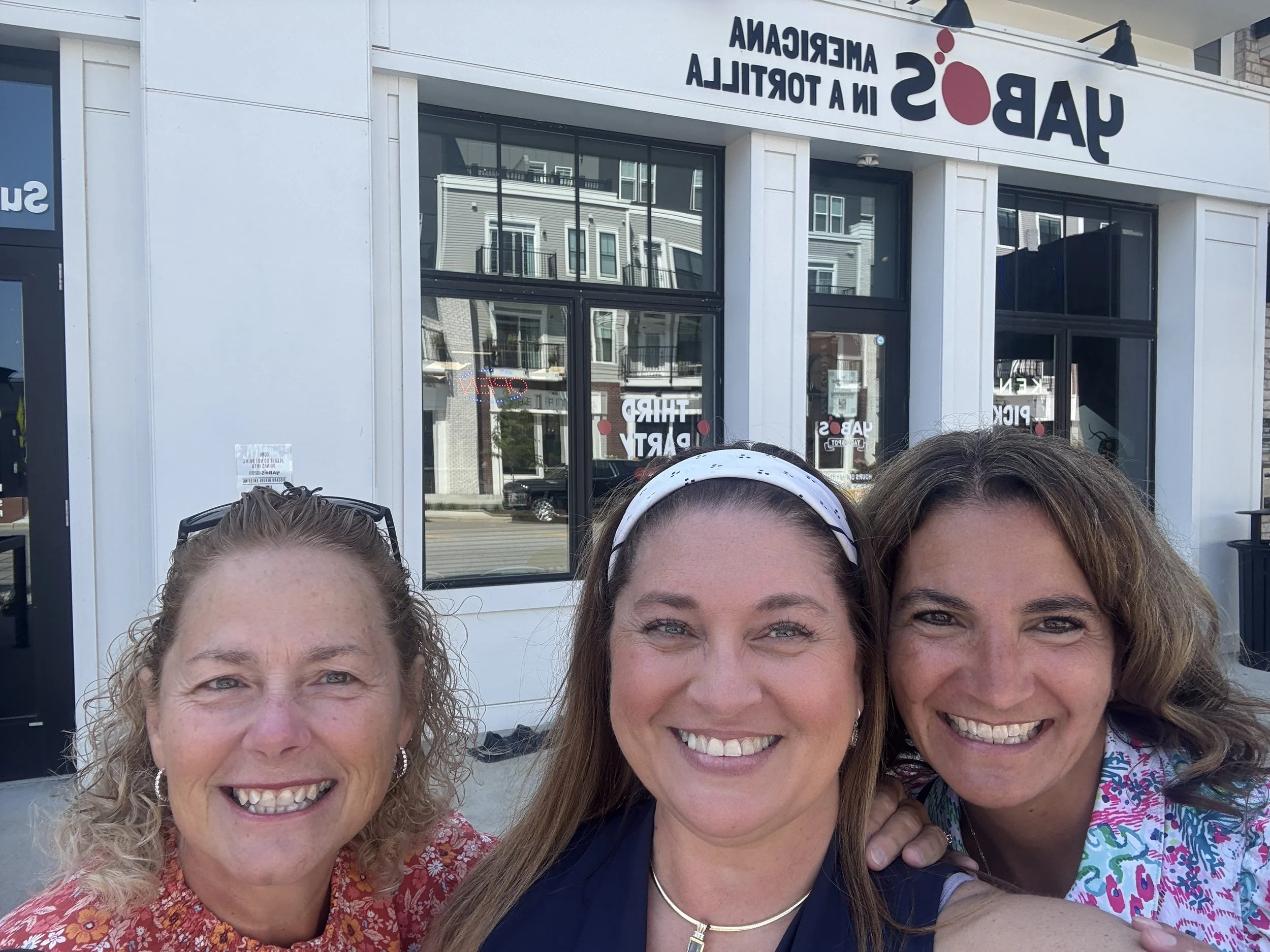 Three women smiling and taking a selfie outside a storefront with a sign that reads "UABOS IN A TERMICAL" in reverse, with buildings reflected in the windows.