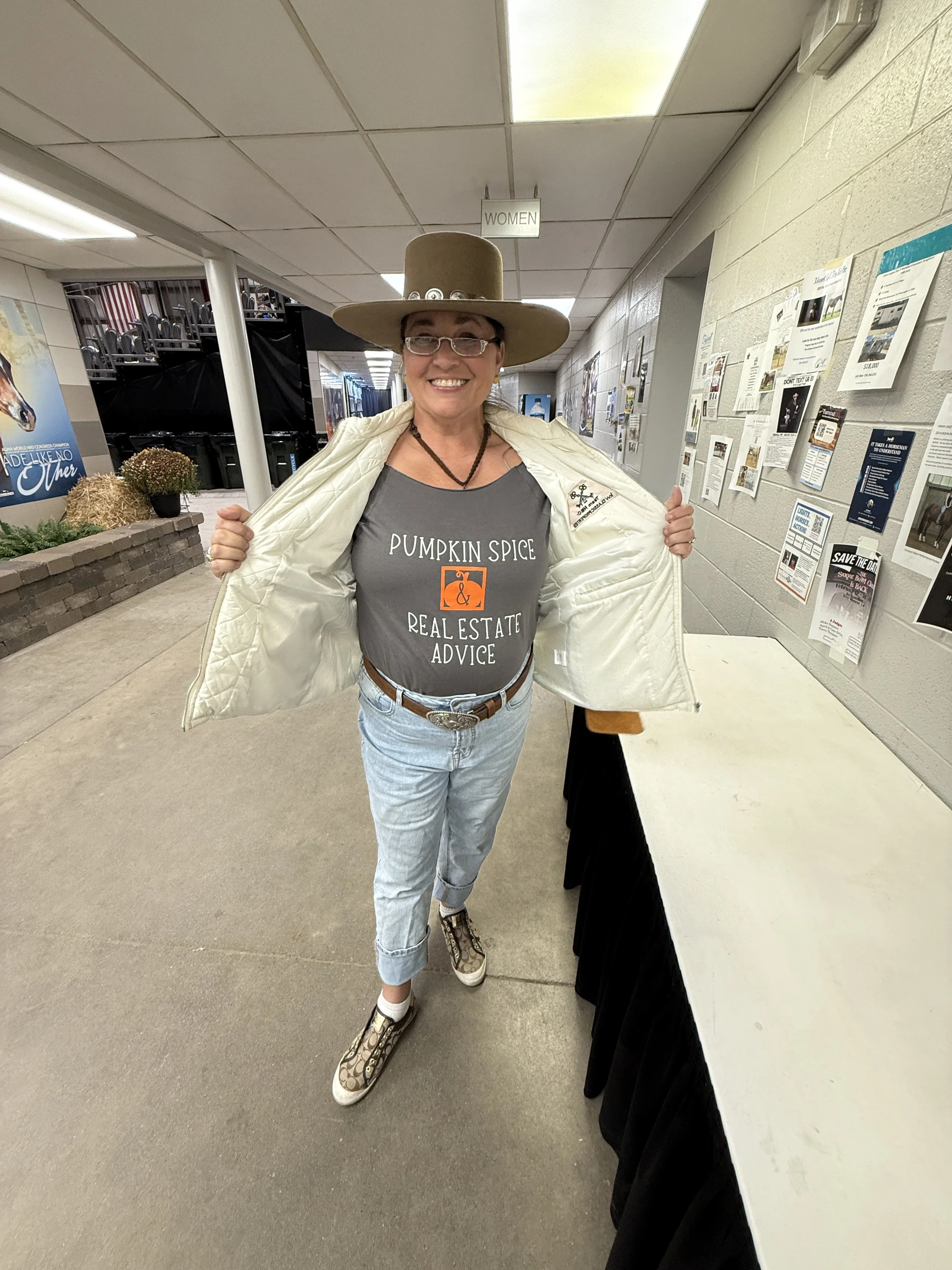 A woman wearing a large tan hat, glasses, a gray T-shirt with text, a white jacket, light blue jeans, and patterned sneakers, standing in a hallway with bulletin boards and a women’s restroom sign overhead.