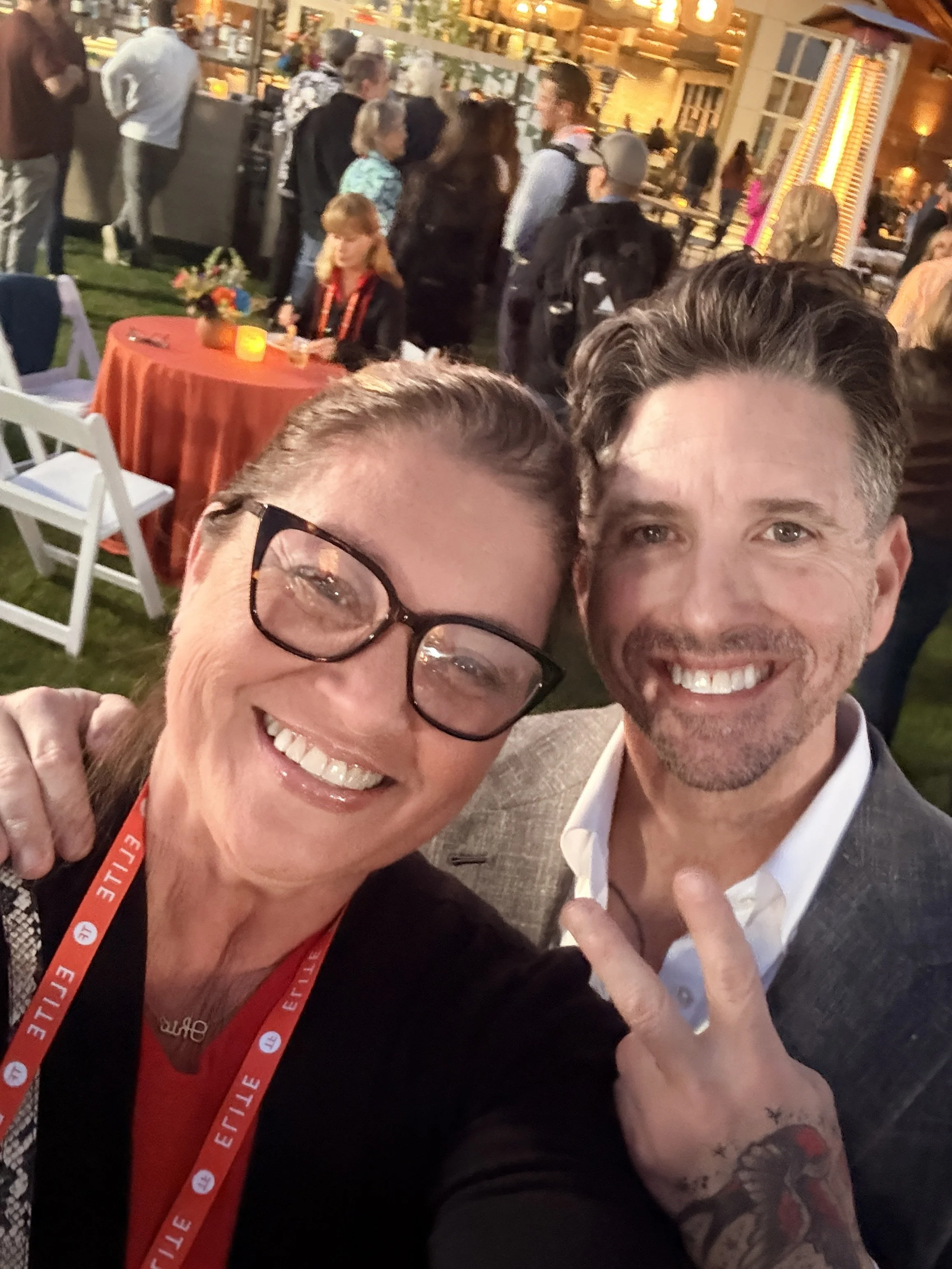 Close-up of a woman and a man smiling at a social event with many people in the background. The woman is wearing glasses and a red lanyard, and the man has angels tattoos on his arm and is making a peace sign.
