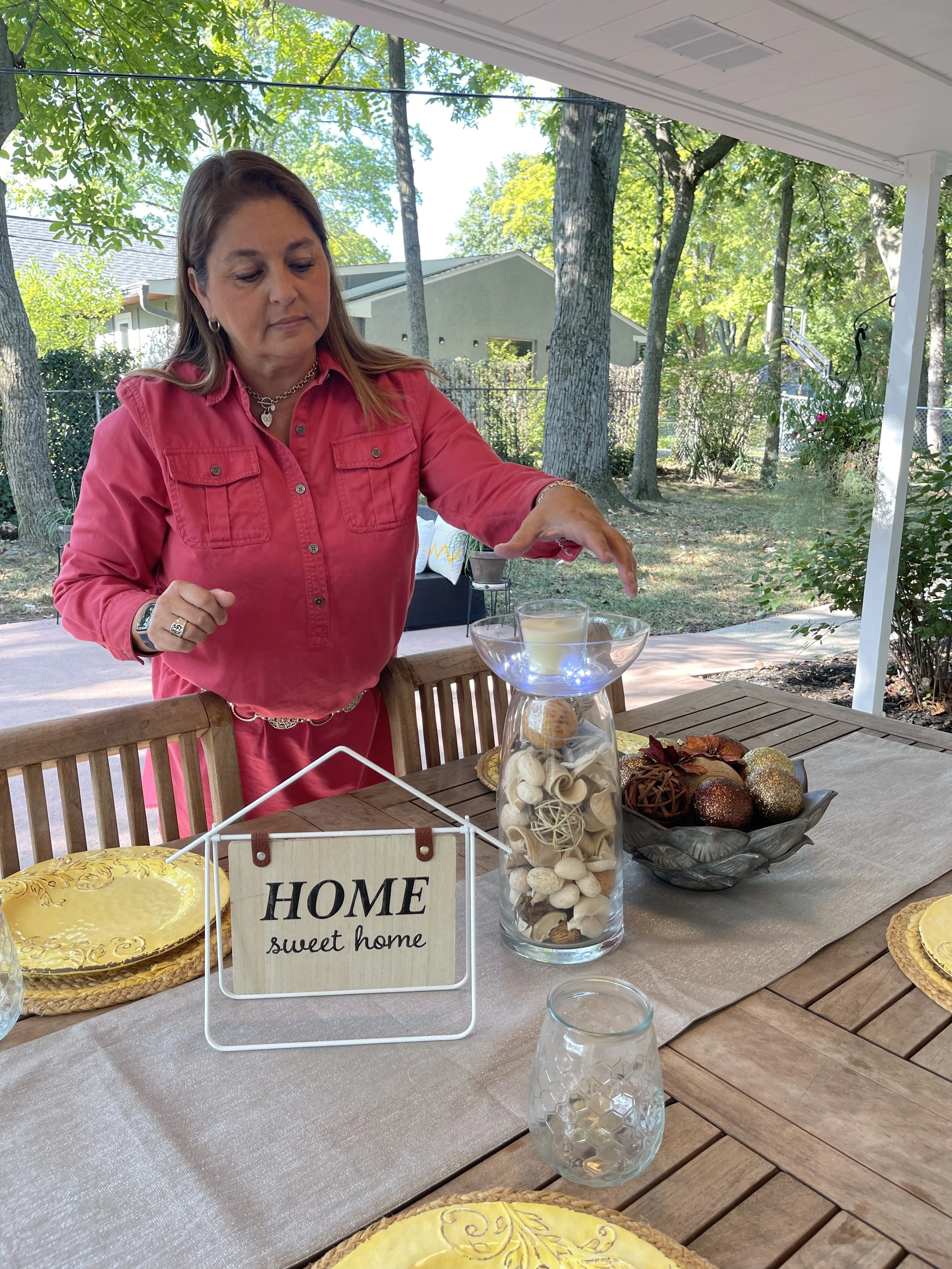 A woman in a pink shirt standing at an outdoor table with decorative fall and Christmas ornaments, a glass bowl with a candle, and a small "Home sweet home" sign. The table is covered with a beige tablecloth and has empty glasses. The background includes trees, a house, and a yard.