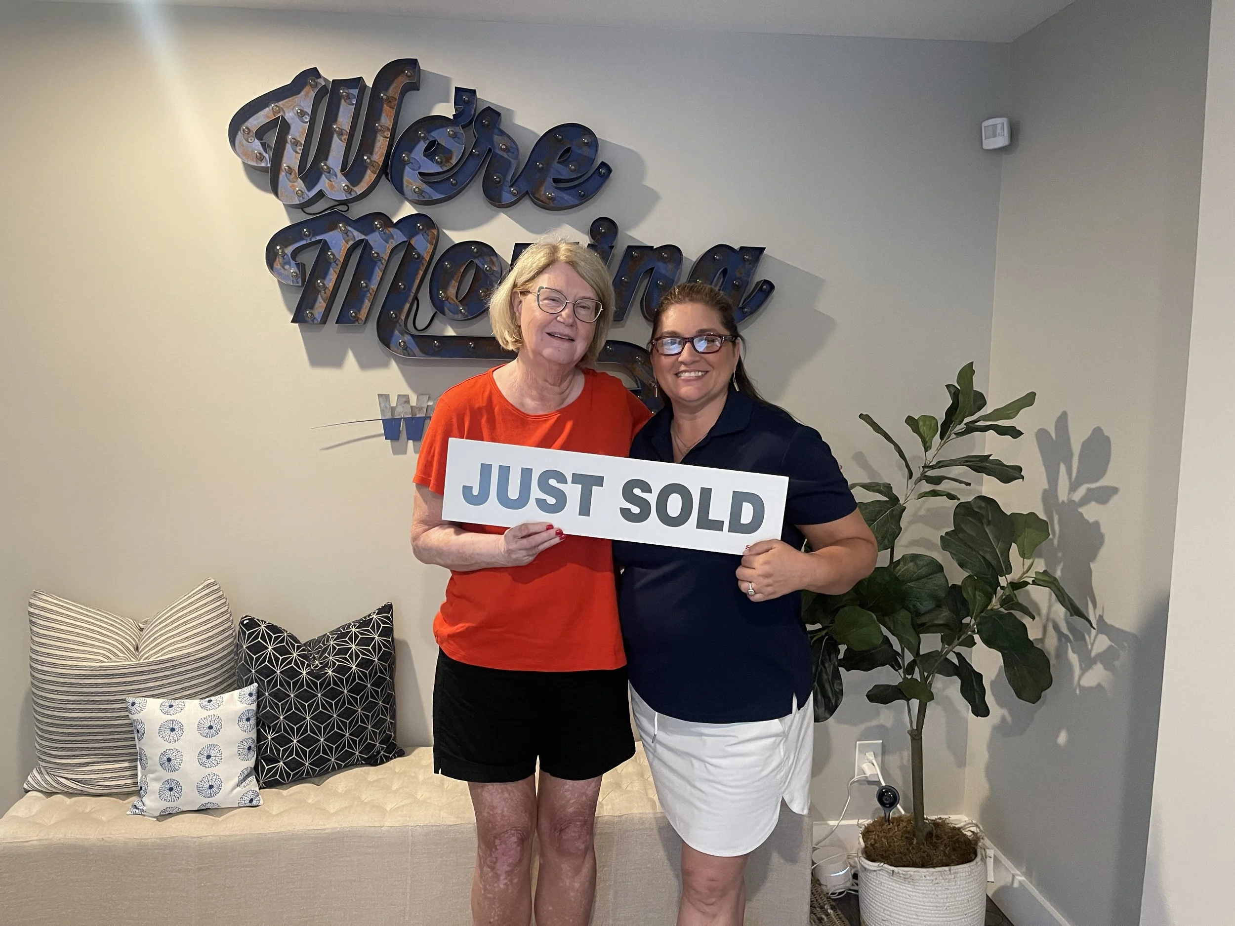 Two women standing in front of a wall with a decorative sign reading 'Wren & Moriah.' One woman is holding a sign that says 'JUST SOLD.' They are smiling, and there are patterned pillows on a bench behind them, along with a large houseplant on the ri