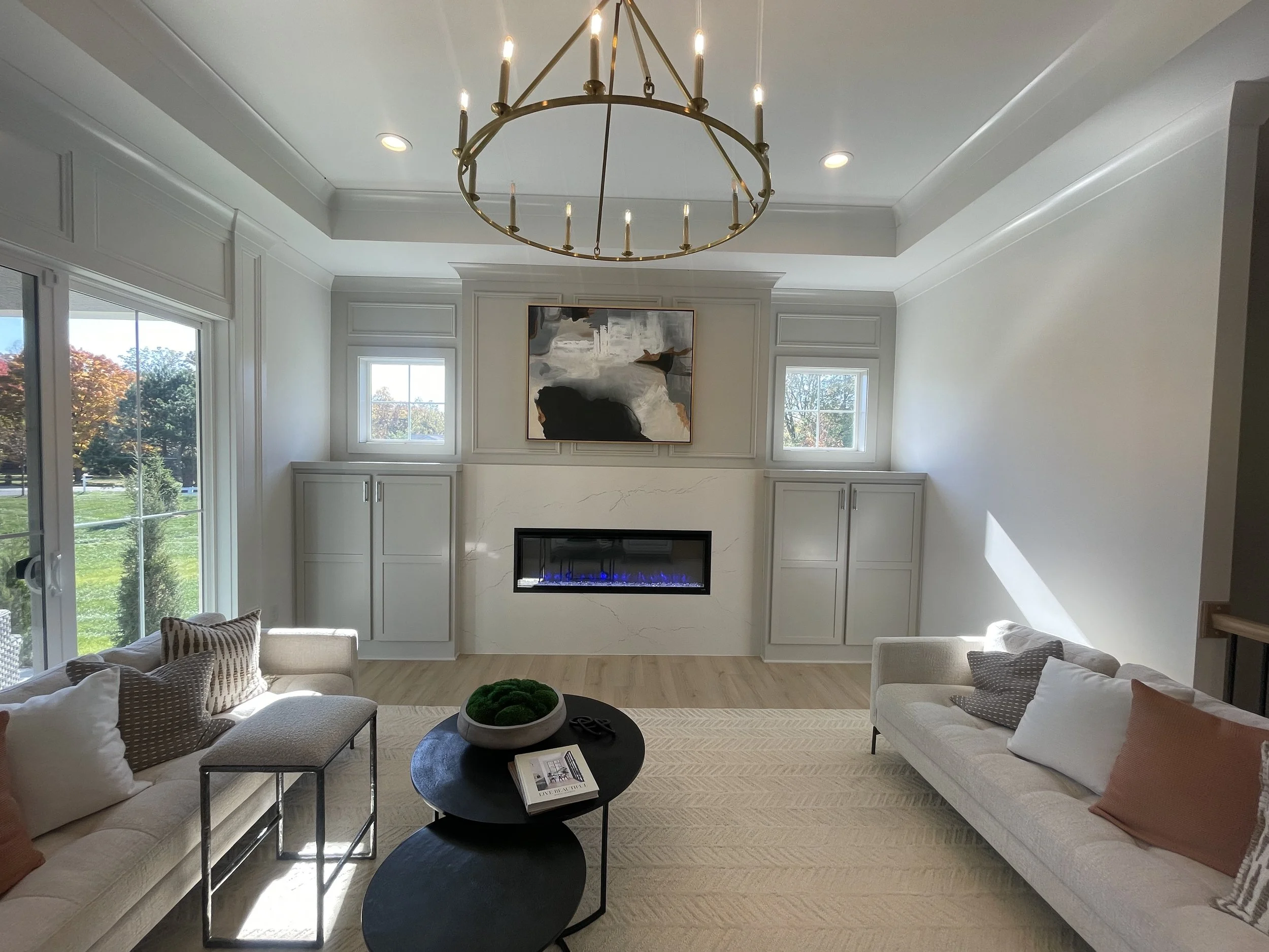 Living room with white walls, a fireplace, large windows showing trees outside, and modern furniture including sofas, a coffee table with a book and decor, and a chandelier hanging from the ceiling.