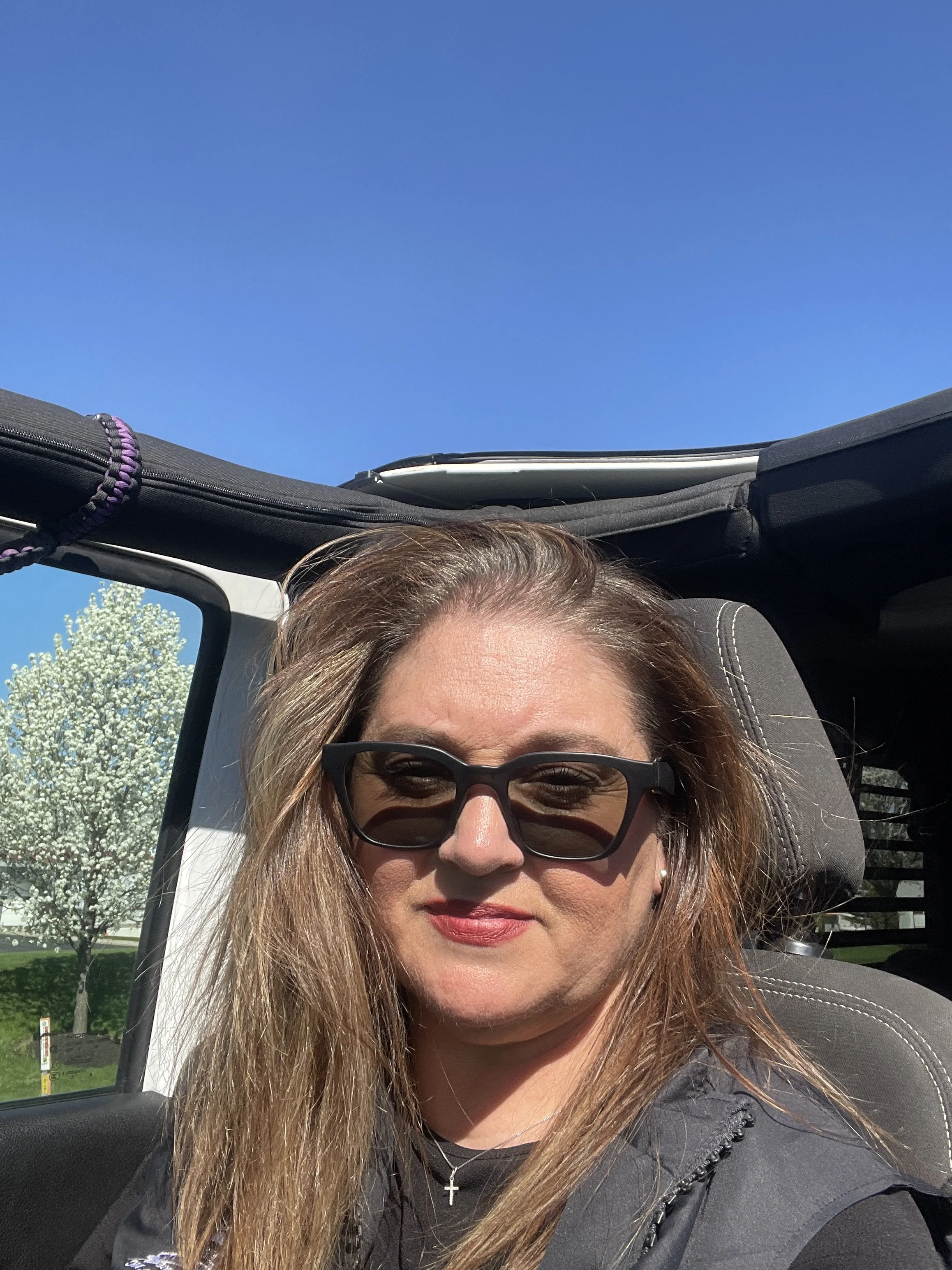 Close-up of a woman with long brown hair wearing sunglasses, a black jacket, a necklace with a cross, sitting in a car with the top down, under a clear blue sky with white blooming trees in the background.
