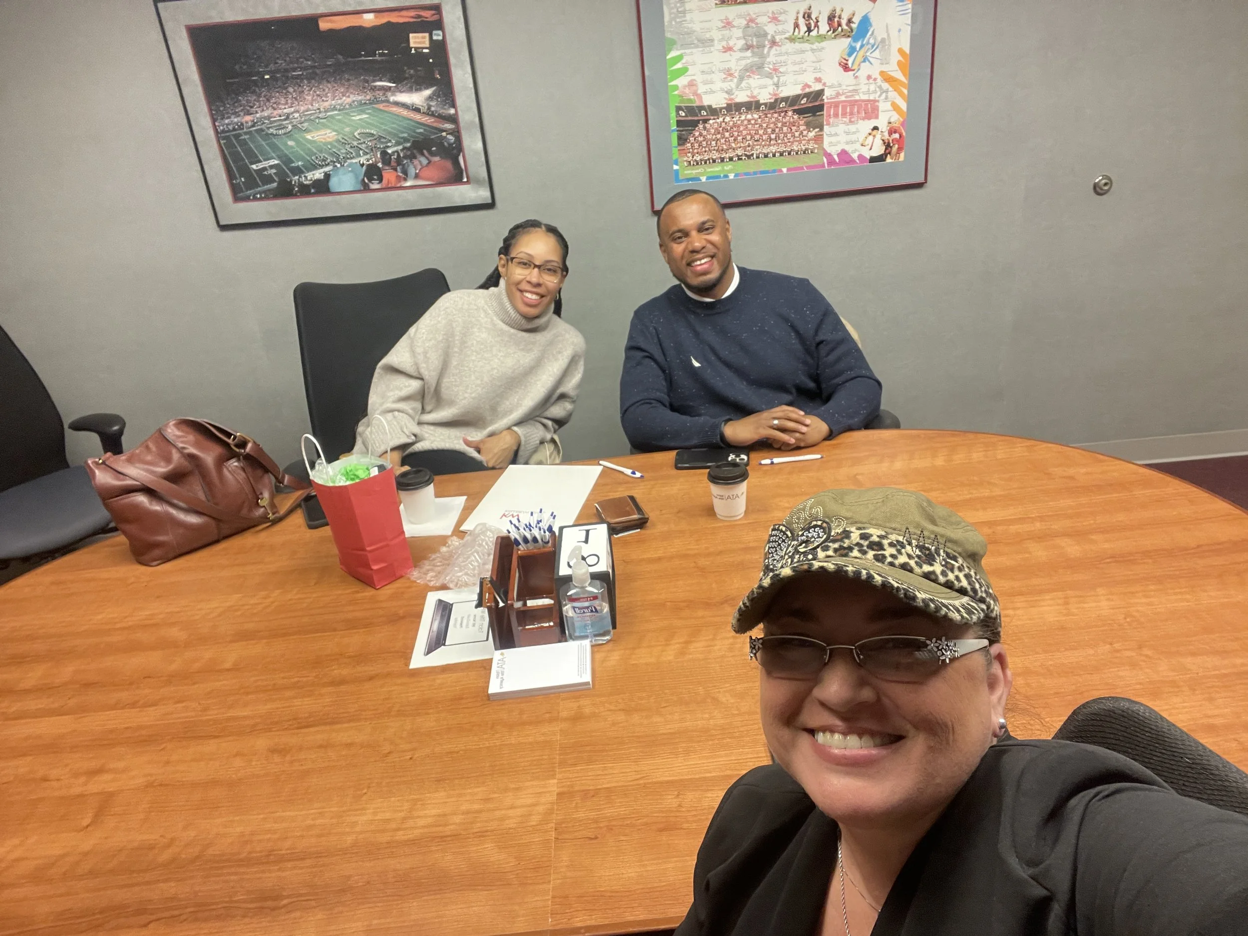 A woman taking a selfie with three people sitting at a conference table behind her; two women and one man smiling and looking at the camera. The table has various items including cups, papers, and a gift bag, with sports team photos on the wall behin