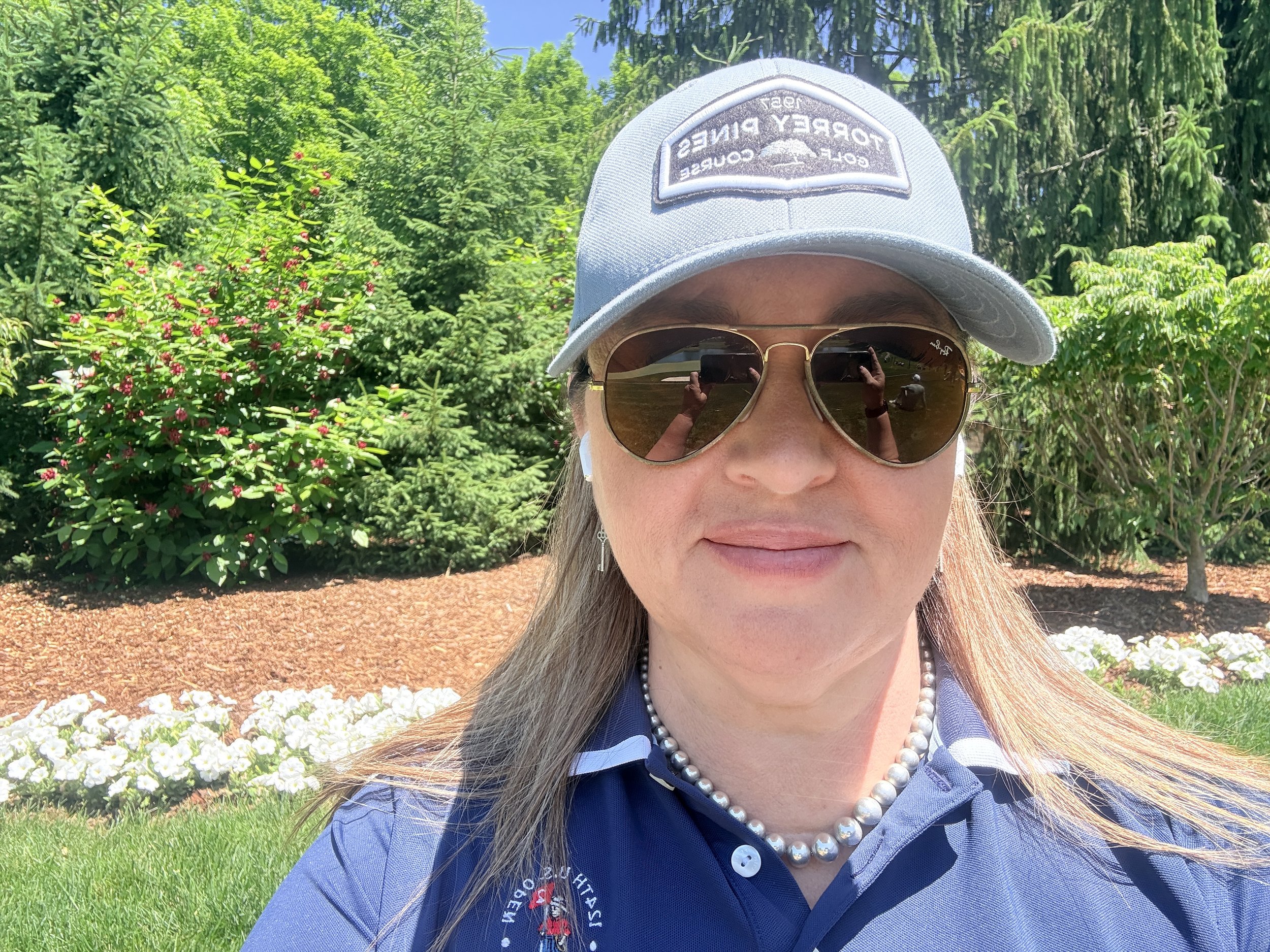A woman wearing sunglasses, a light gray baseball cap, a pearl necklace, and a navy blue polo shirt is outdoors in a garden with lush green trees, bushes, and colorful flowers.