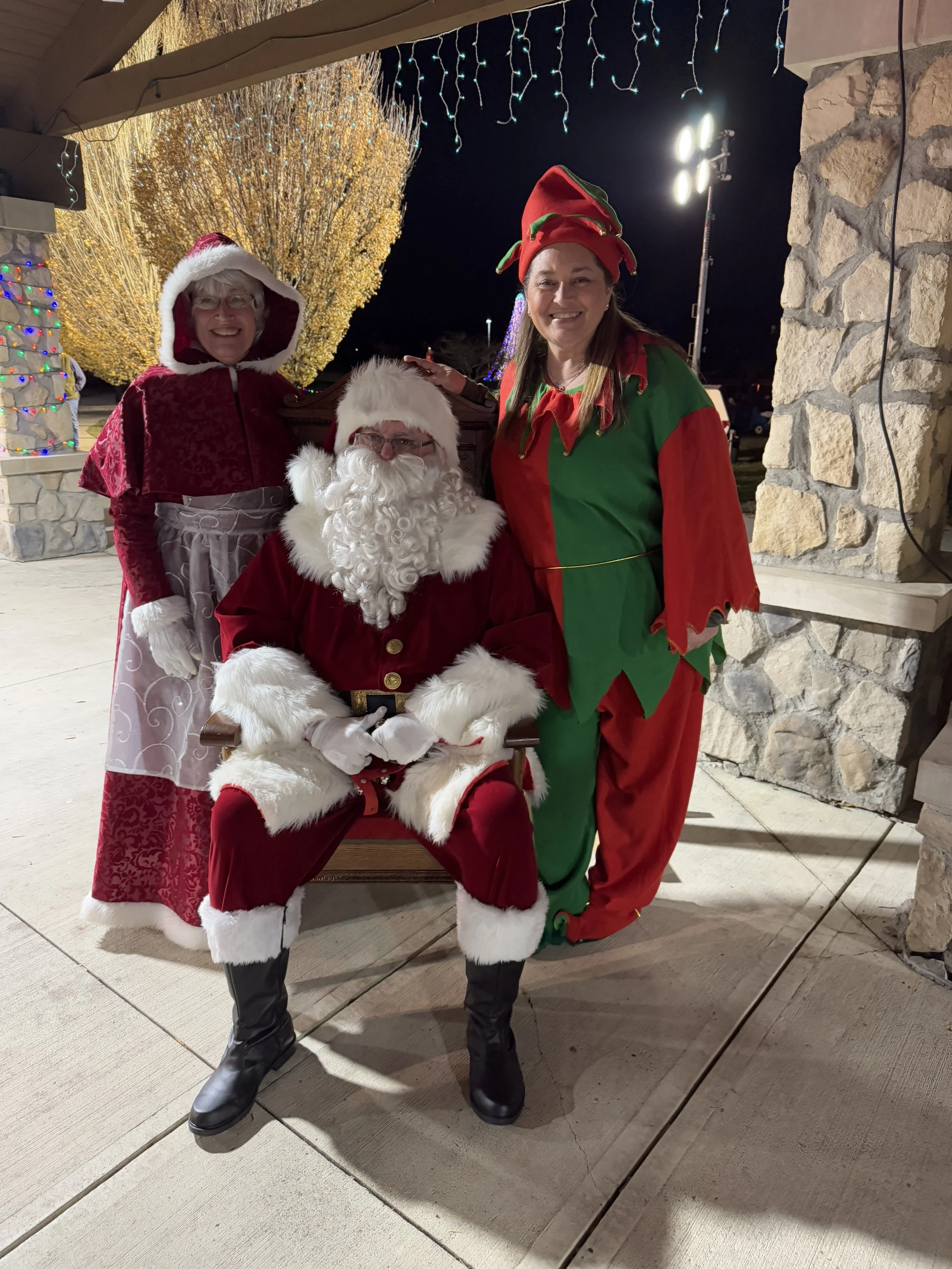 Three people dressed in Christmas costumes posing outdoors at night with Christmas lights and decorated trees in the background.
