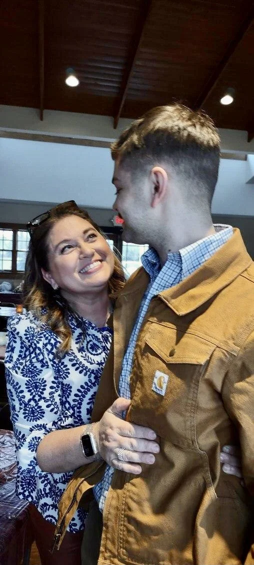 A woman and a young man are smiling and looking at each other in a warmly lit indoor setting, possibly a restaurant or cafe.