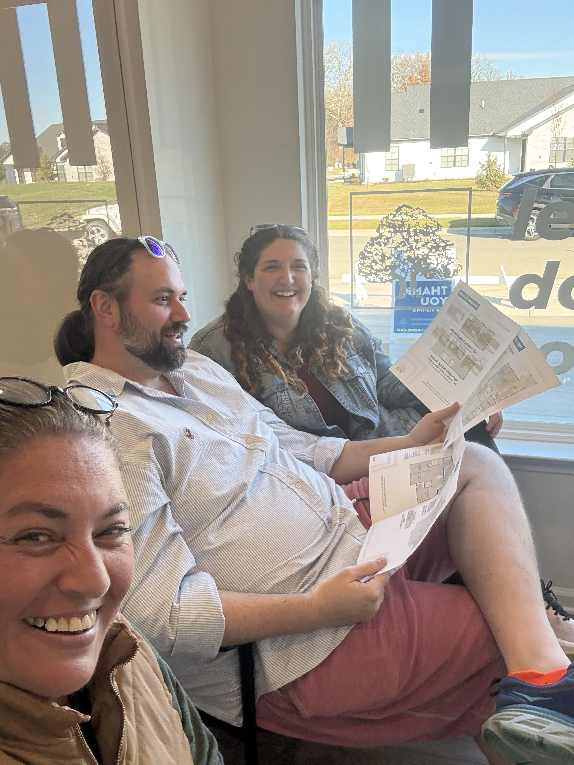 Four people sitting inside a restaurant next to a window, smiling and holding menus, with cars and houses visible outside.