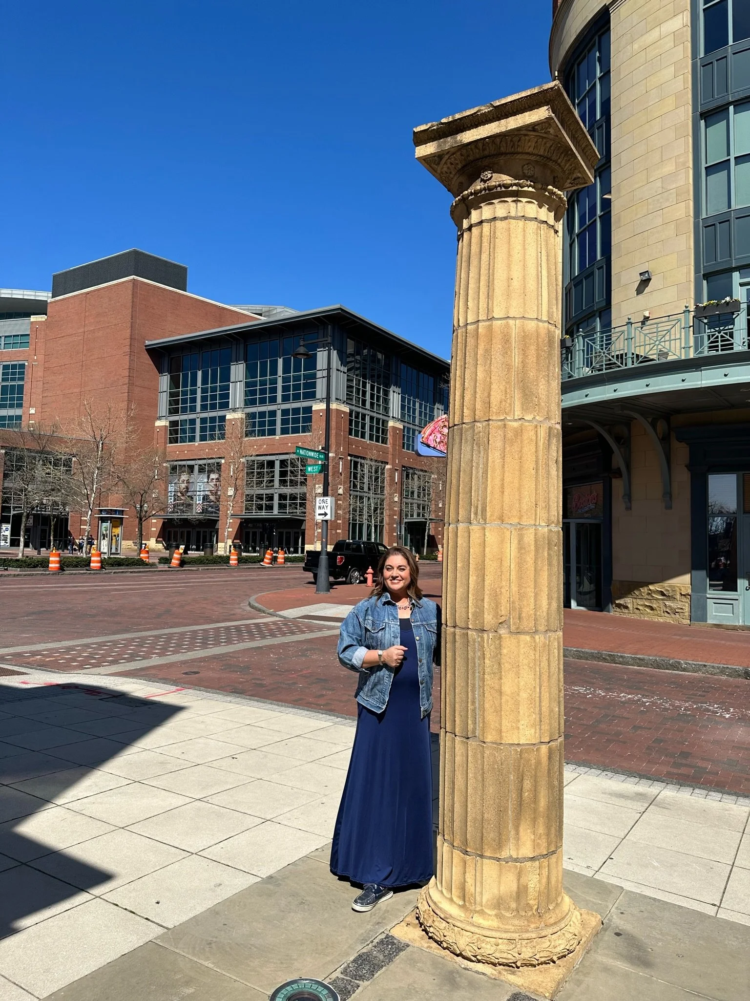 A woman in a denim jacket and dark blue dress standing on a city sidewalk behind a large, ancient-looking stone pillar with carved details. Modern buildings are visible in the background under a clear blue sky.