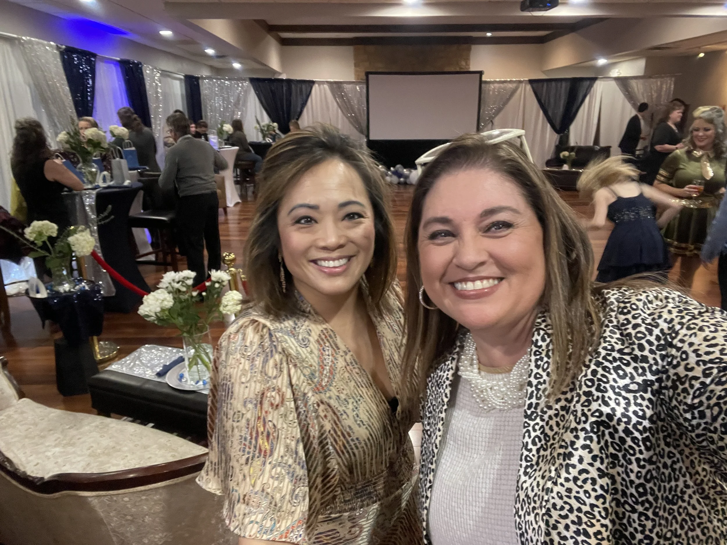 Two women smiling and taking a selfie at a decorated event with tables, flowers, and a projector screen in the background.