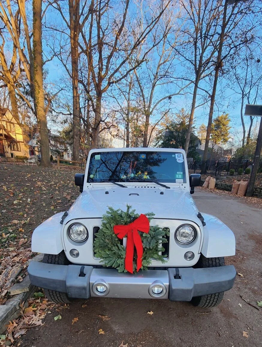 White Jeep with a holiday wreath decorated with a red bow hanging on the front grille, parked on a dirt driveway surrounded by trees with fallen leaves.