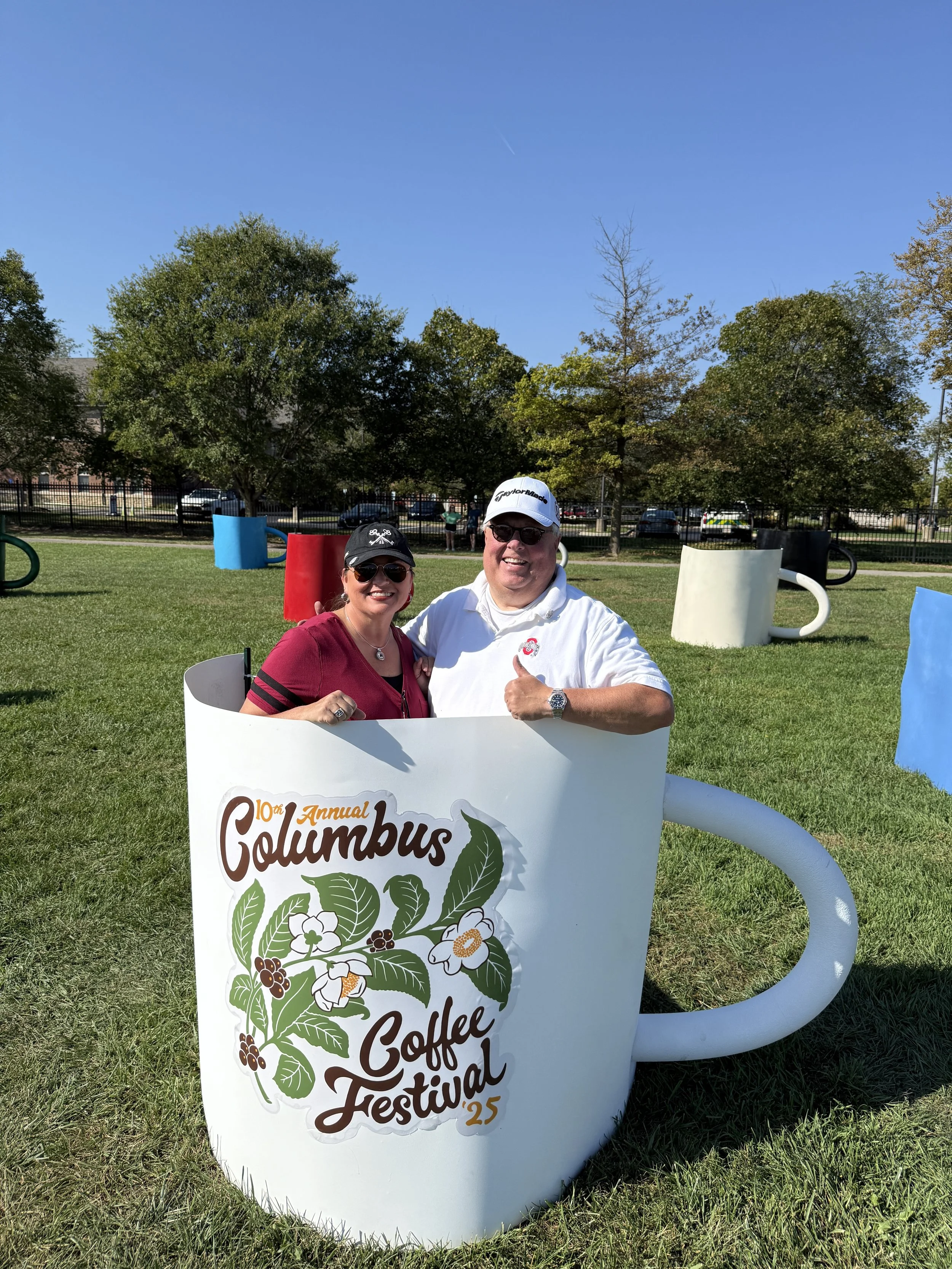 Two people sitting inside a large cup-shaped decoration at a coffee festival outdoors on a sunny day.