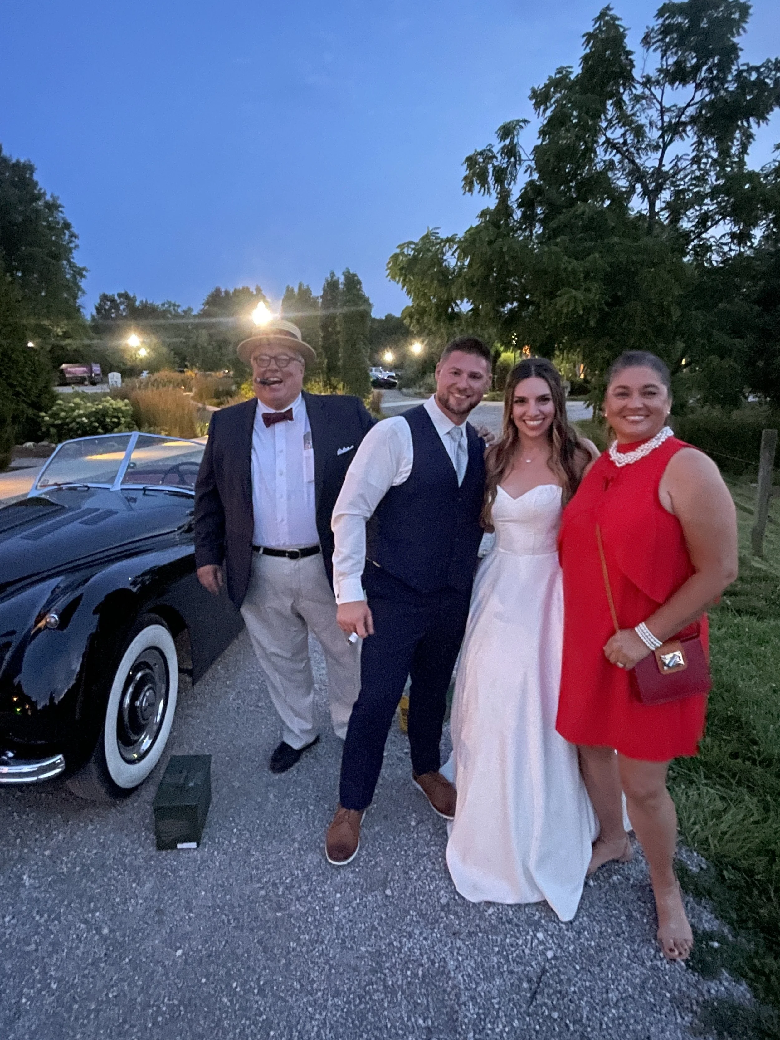 Four people standing outdoors during evening, dressed in formal attire, with a classic black car behind them and trees in the background.