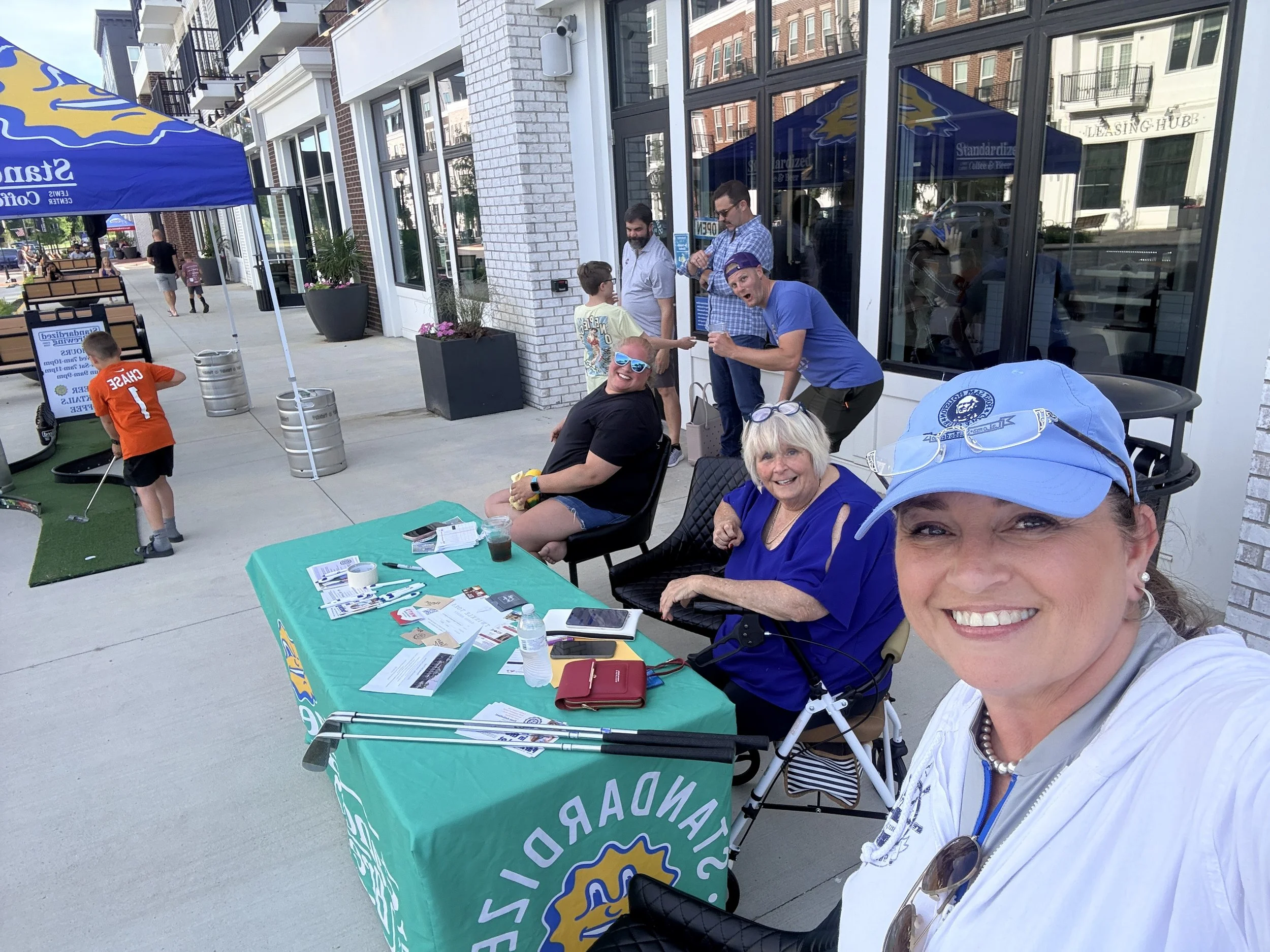 People gathered around a table with promotional materials at an outdoor event, with a sidewalk, buildings, and a blue and yellow tent in the background.