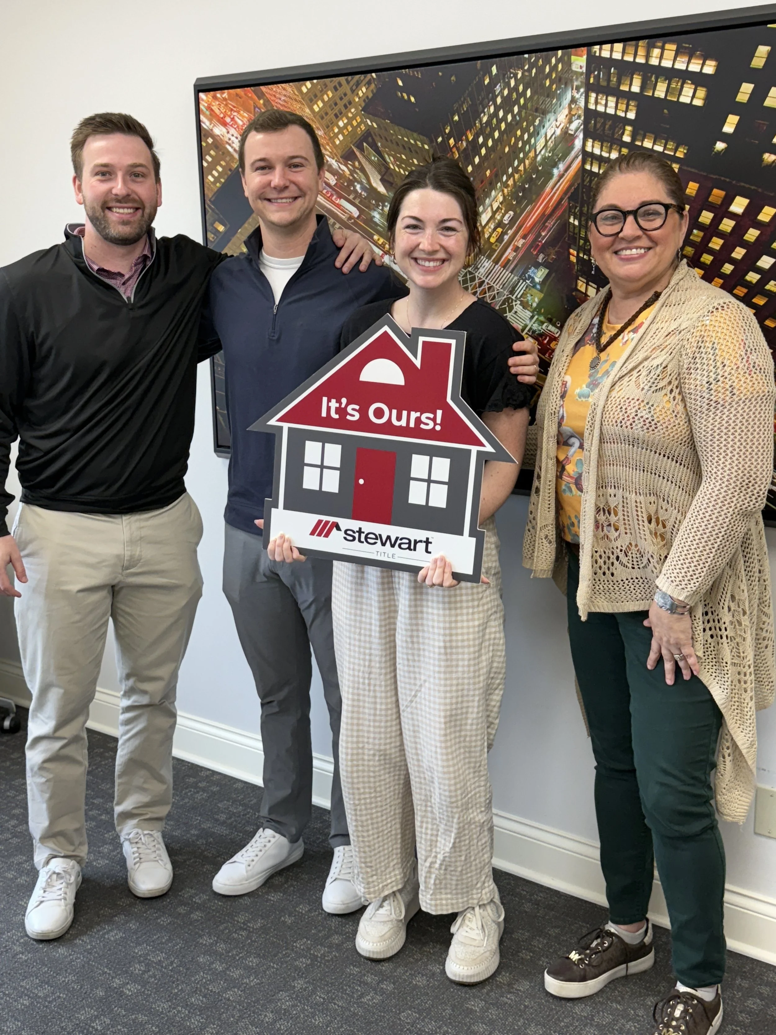 Four people standing together indoors, smiling, with a cityscape photo on the wall behind them. The woman in the center is holding a sign that says, 'It's Ours!'