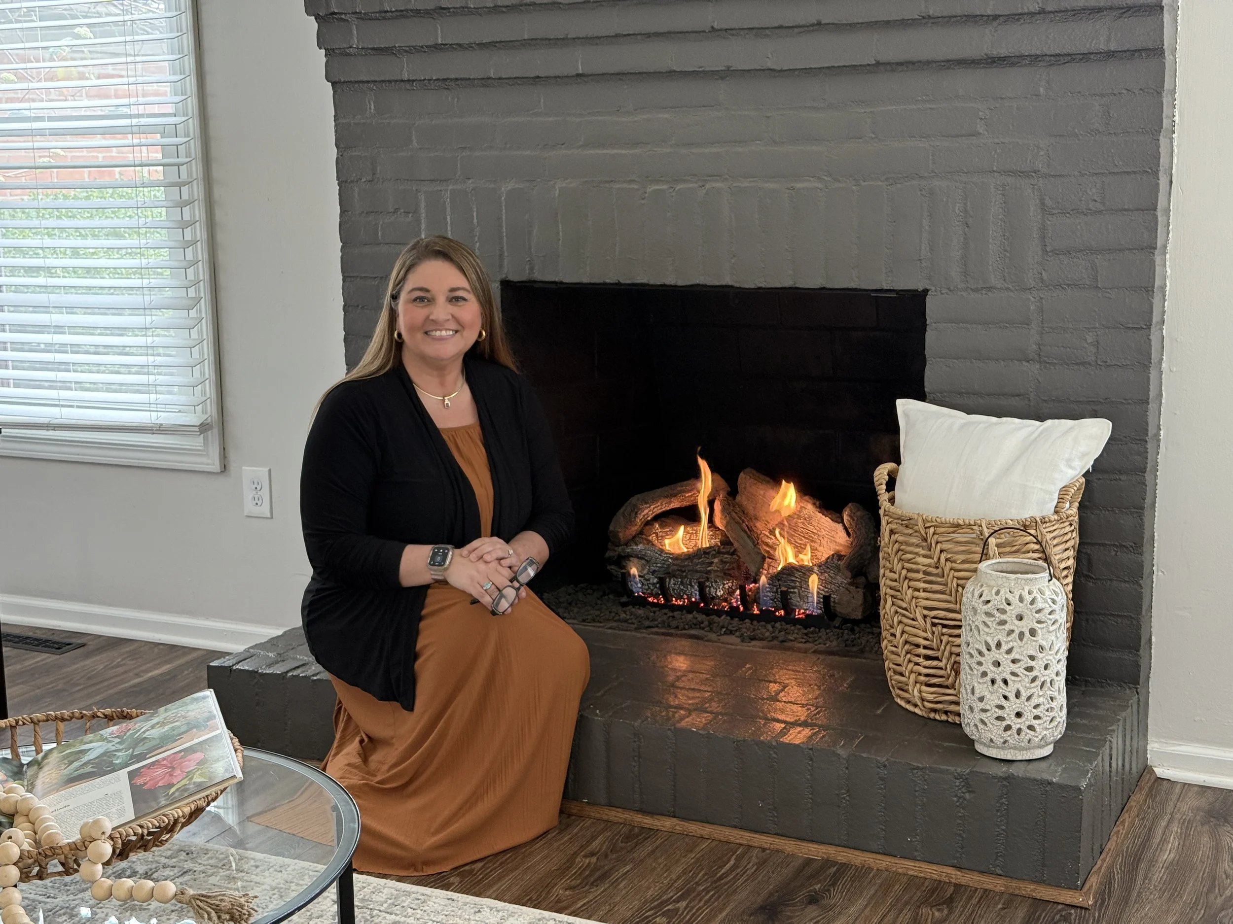 A woman sitting by a fireplace with flames. She is smiling, wearing a black top, gold jewelry, and an orange-brown skirt. The room has a large window with blinds, decorative vases, and a glass table with magazines.