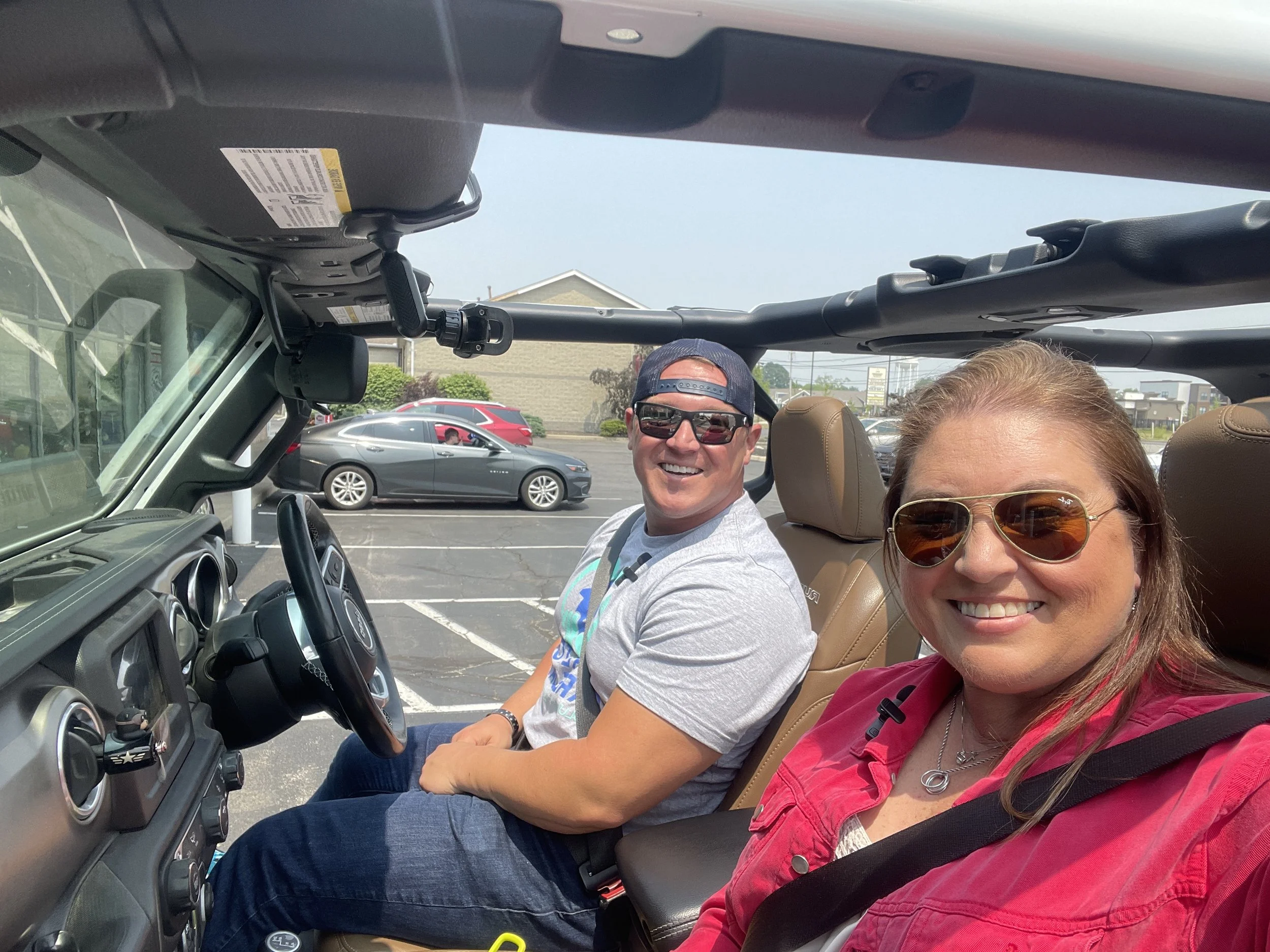Two women and a man inside a convertible vehicle parked in a parking lot. The woman in front is wearing sunglasses and a pink jacket. The man next to her is wearing sunglasses, a gray T-shirt, and a blue cap worn backwards. The background shows other