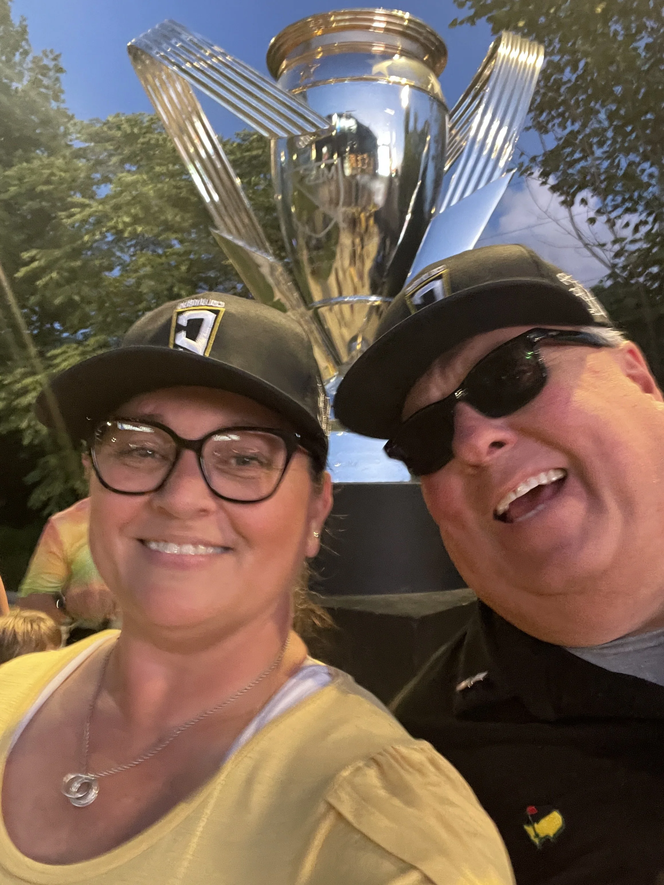 Two women taking a selfie with a large trophy in the background during the evening.