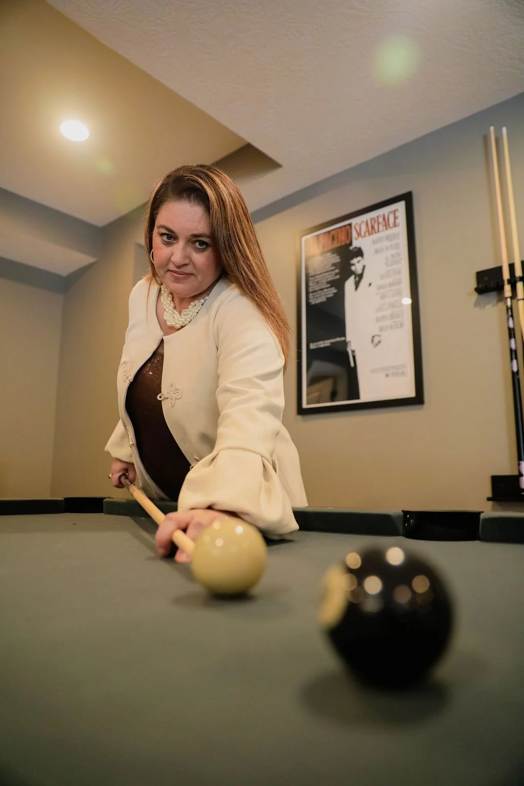A woman playing pool, leaning over the table, with a focused expression, holding a pool cue, with a framed movie poster behind her.