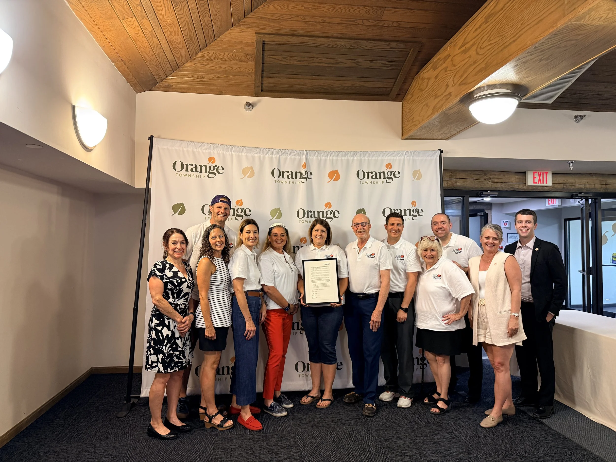 Group of people standing together at an event with a backdrop that reads 'Orange Township'. They are smiling and some are holding a framed certificate. The group includes men and women of different ages dressed in semi-formal and casual attire, stand