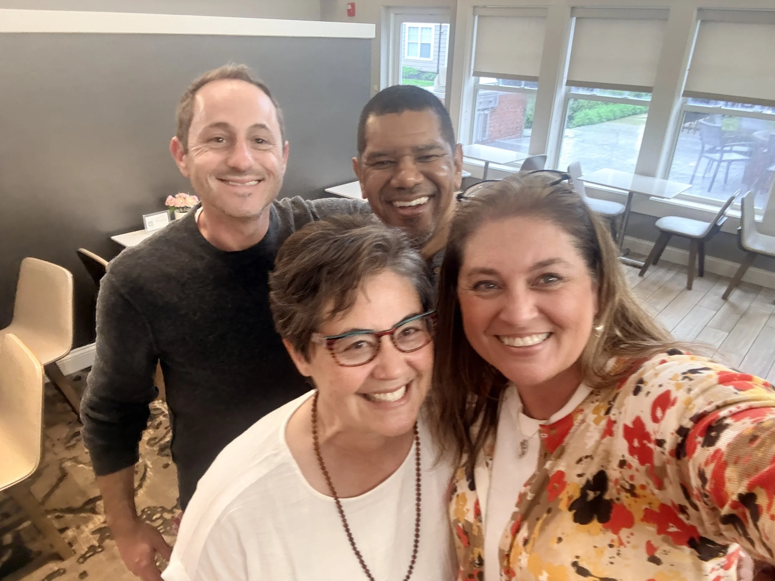 Group of five smiling people taking a selfie in a well-lit room with large windows, tables, and chairs.