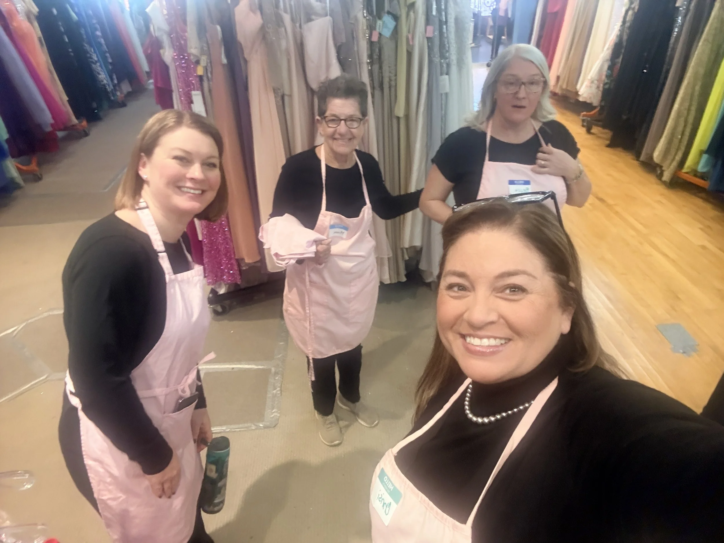 Three women wearing pink aprons in a clothing store surrounded by racks of dresses.