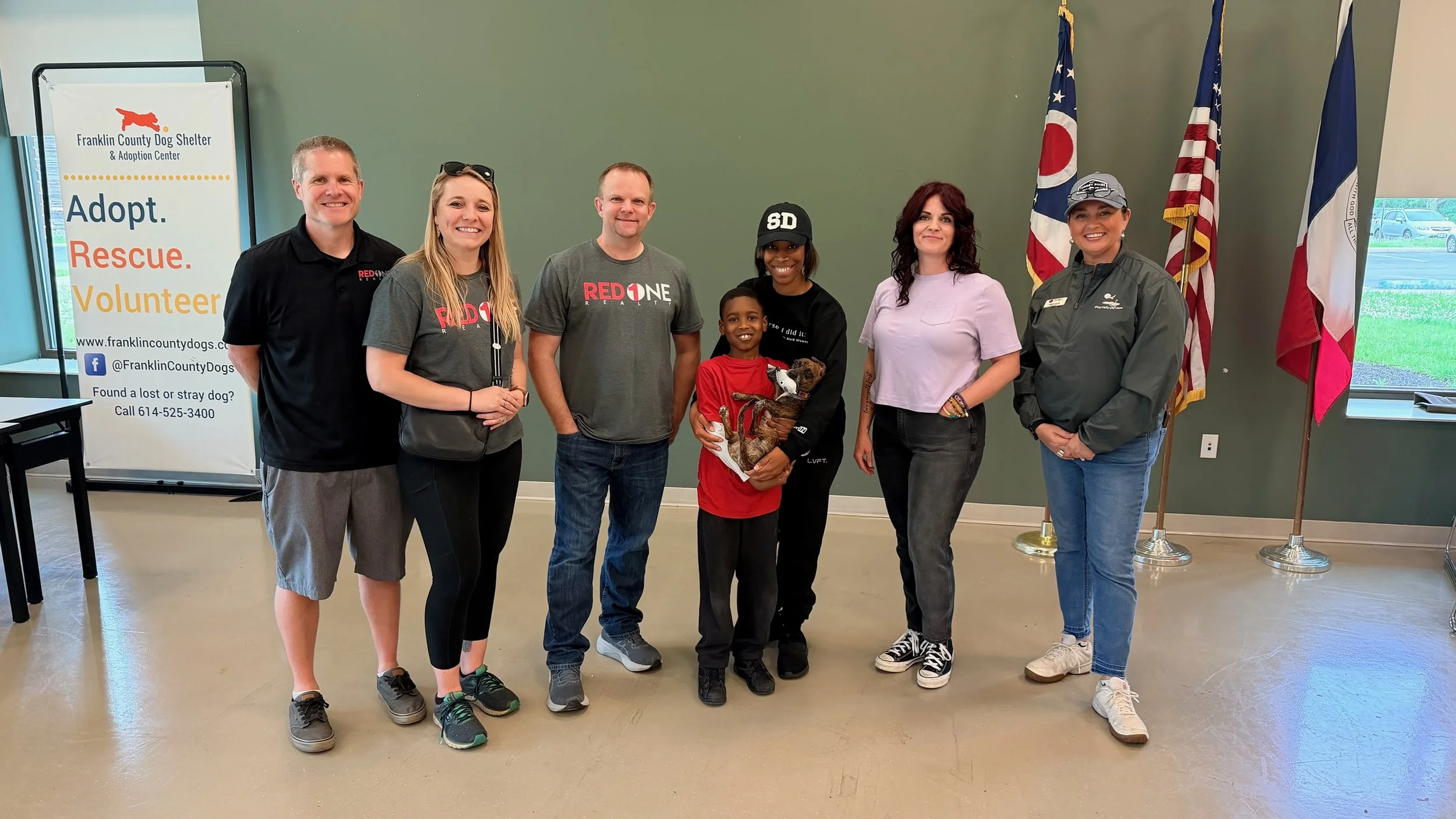 Group of seven people standing inside a room at Franklin County Dog Shelter, with flags and a sign promoting adoption and rescue of dogs in the background.
