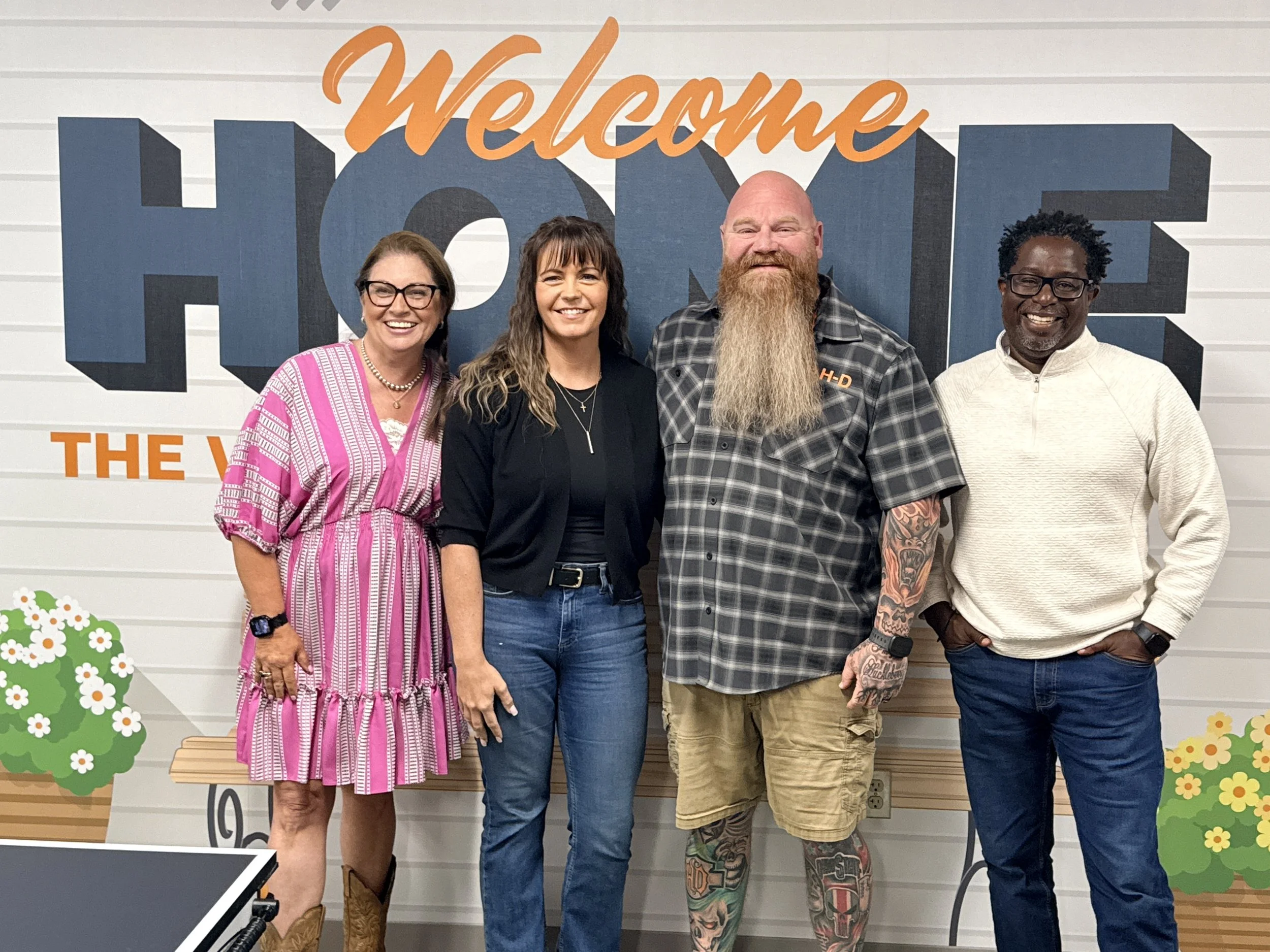 Four people standing in front of a wall with a 'Welcome Home' sign, smiling for the camera. The wall has cartoon-like trees and flowers at the bottom corners.