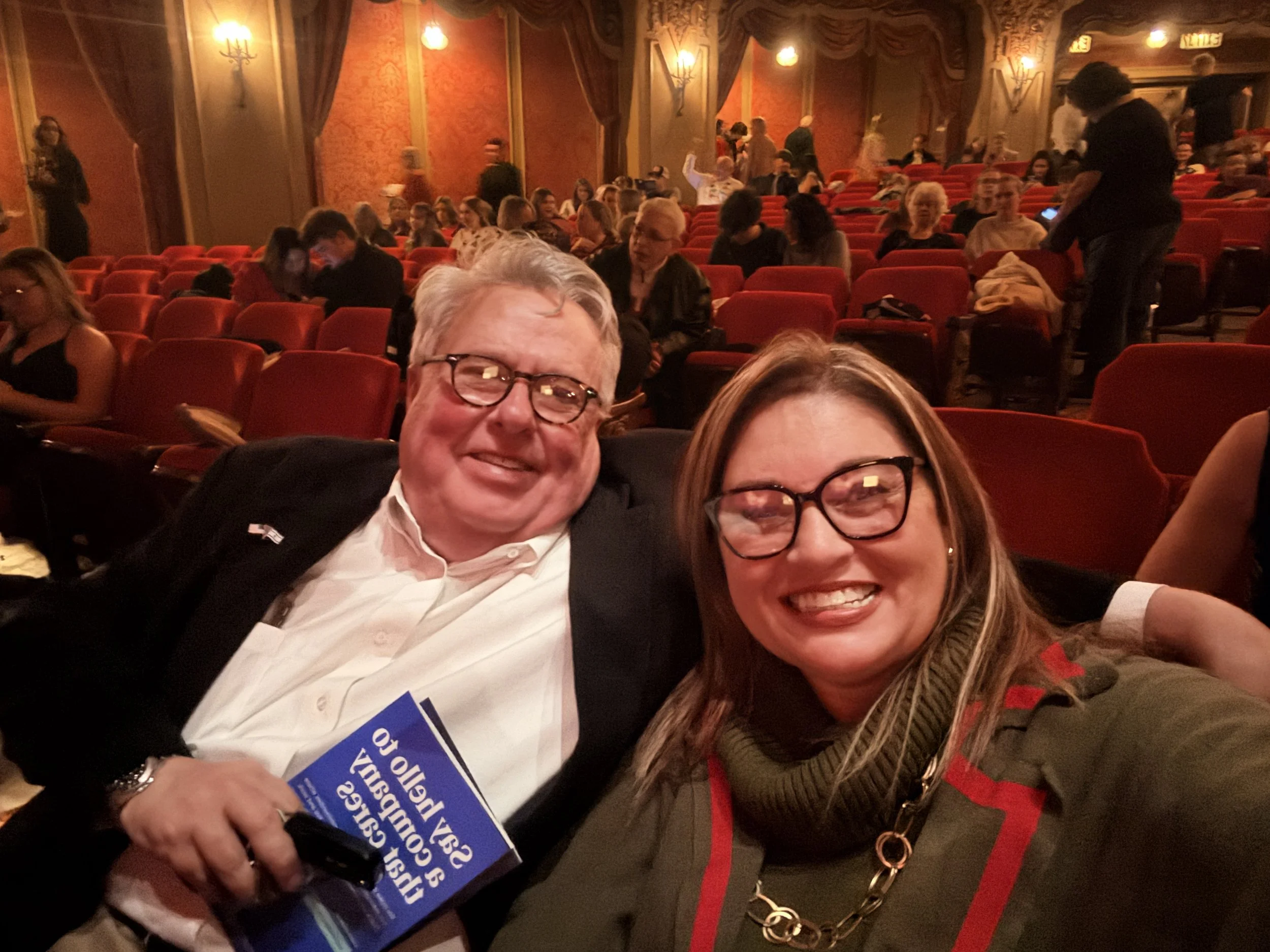 Two smiling people, a man and a woman, taking a selfie in a crowded theater with red seats and ornate decor on the walls, holding a blue campaign sign.
