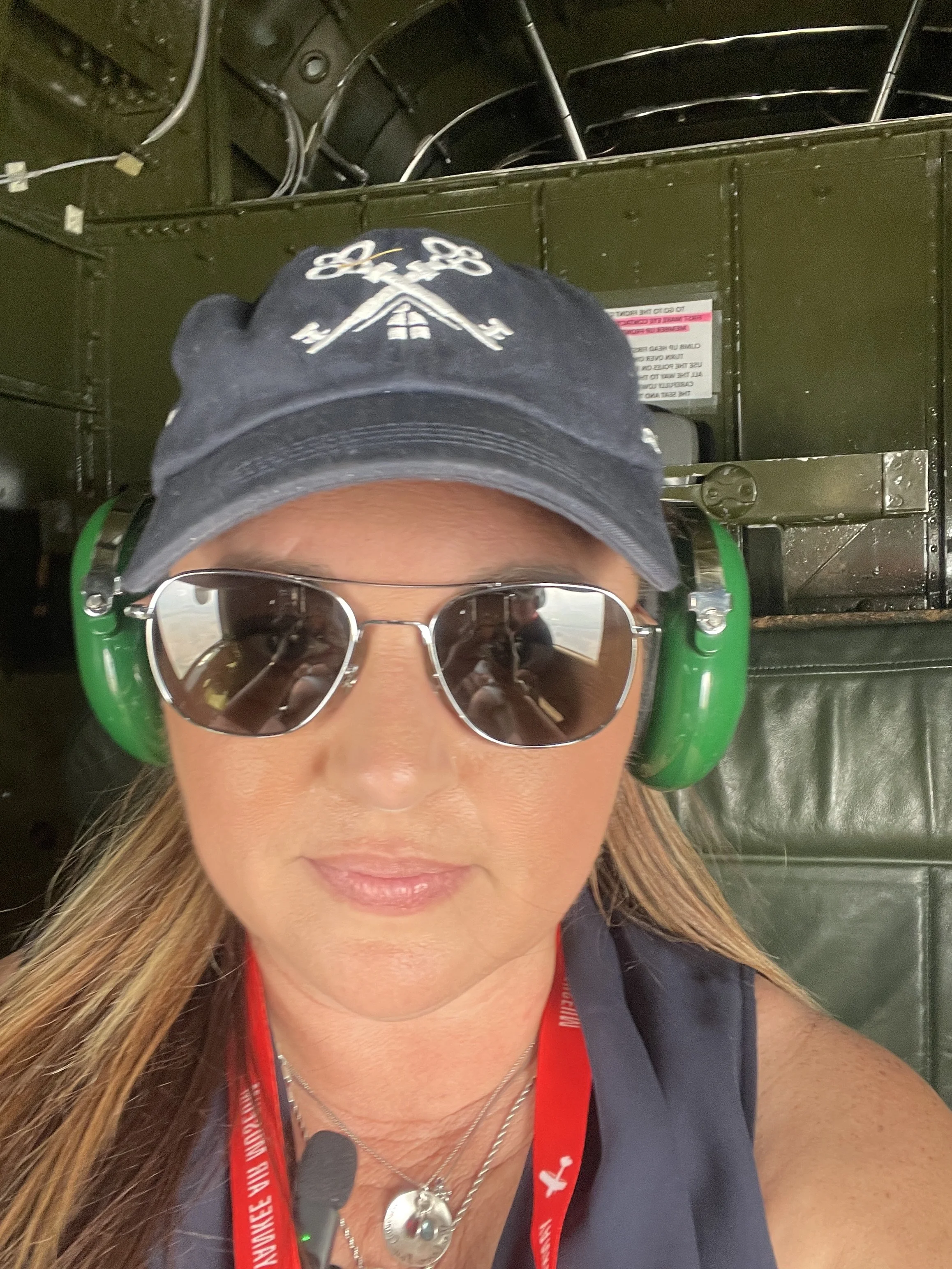 A woman with long hair wearing sunglasses, a black cap with scissors and house symbols, a red lanyard, and headphones, standing in front of a military aircraft interior.
