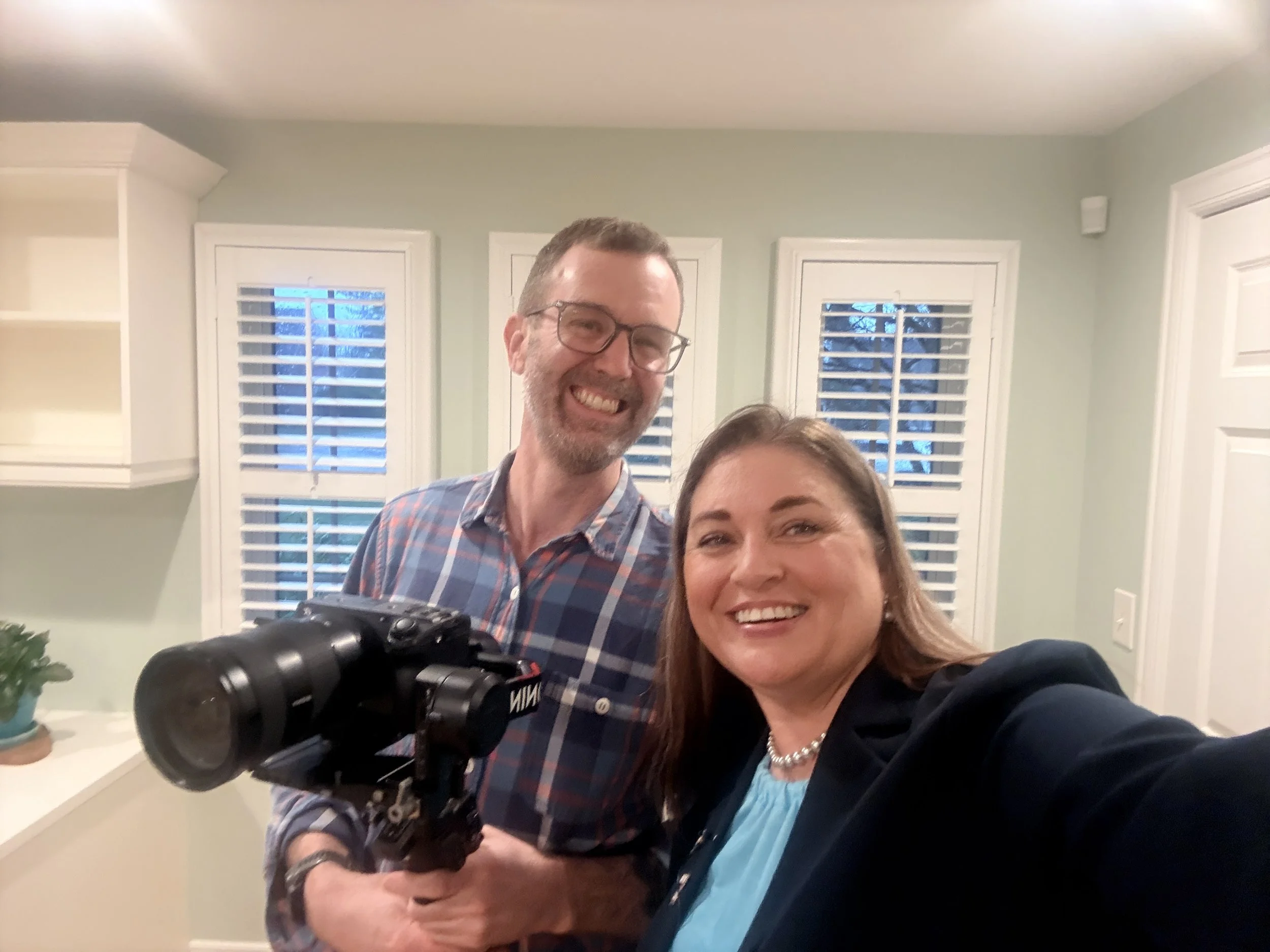 Two smiling adults, a man and a woman, taking a selfie in a bright room with green walls and white window blinds. The man has short hair, glasses, and a beard, and is holding a camera. The woman has shoulder-length brown hair and is wearing a pearl n