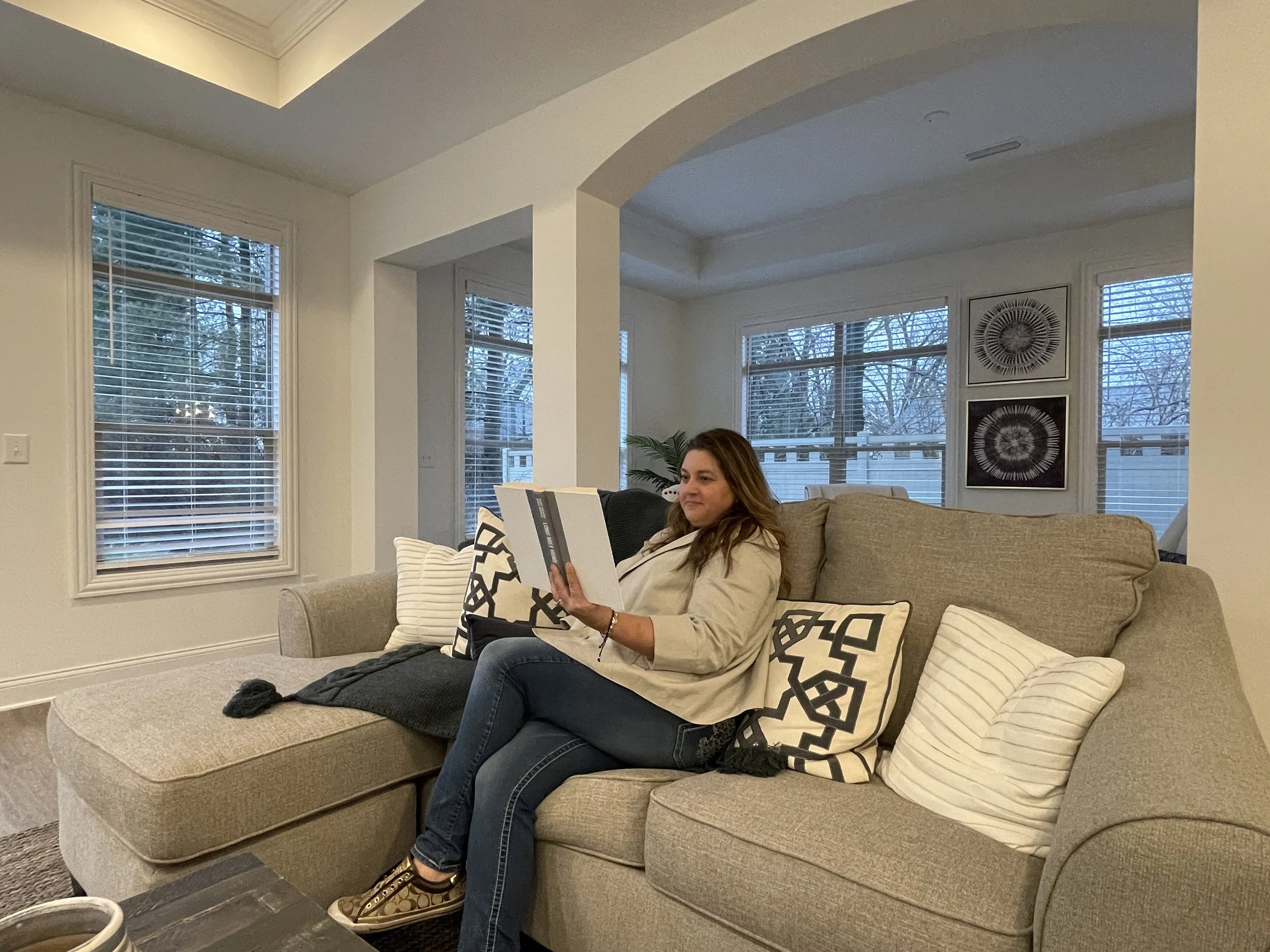 A woman sits on a beige sectional sofa in a bright living room, reading a book. The room has large windows with white blinds, and there are black and white framed artworks on the wall.