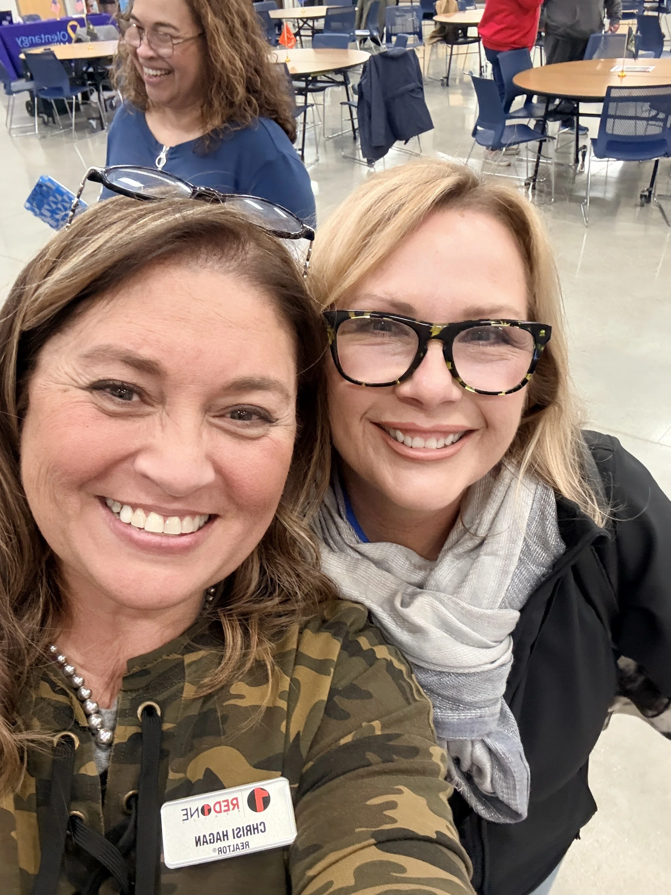 Two smiling women taking a selfie at a voting event, with one woman wearing black and yellow patterned glasses, a gray scarf, and a black jacket, and the other woman wearing a camo jacket and glasses. In the background, a woman with curly brown hair 