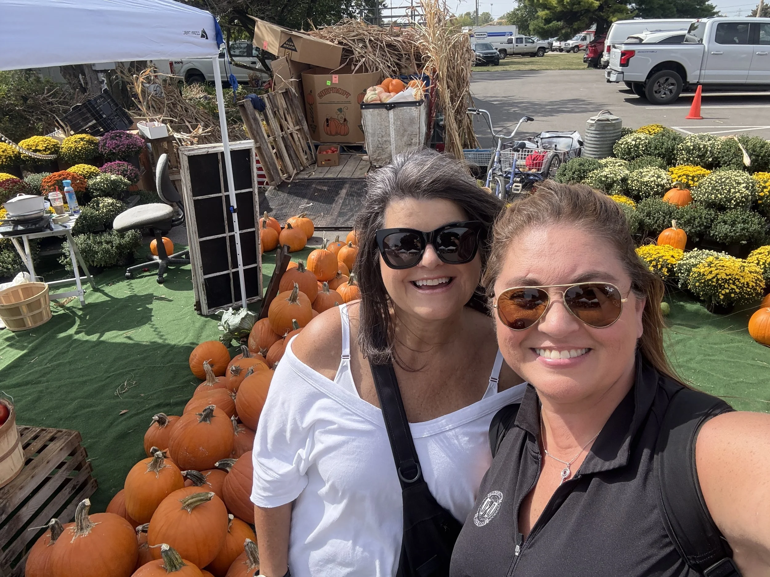 Two women smiling and taking a selfie at a fall market with pumpkins, chrysanthemums, and other fall decorations around them.