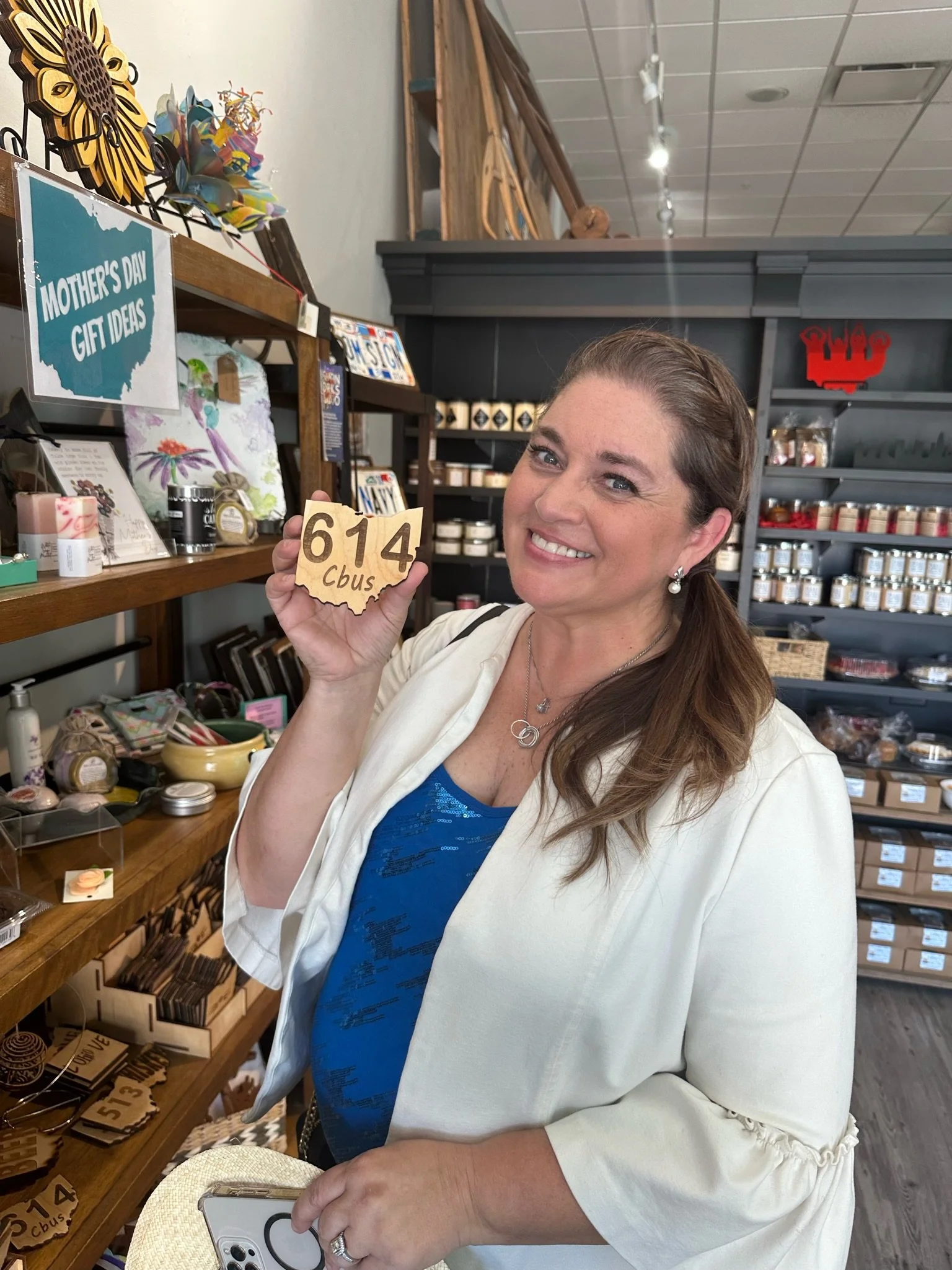 A woman with brown hair smiling and holding a wooden sign that reads '614 Cbus.' She is standing in a store with shelves of assorted items and gift ideas, some with signs indicating they are for Mother's Day.