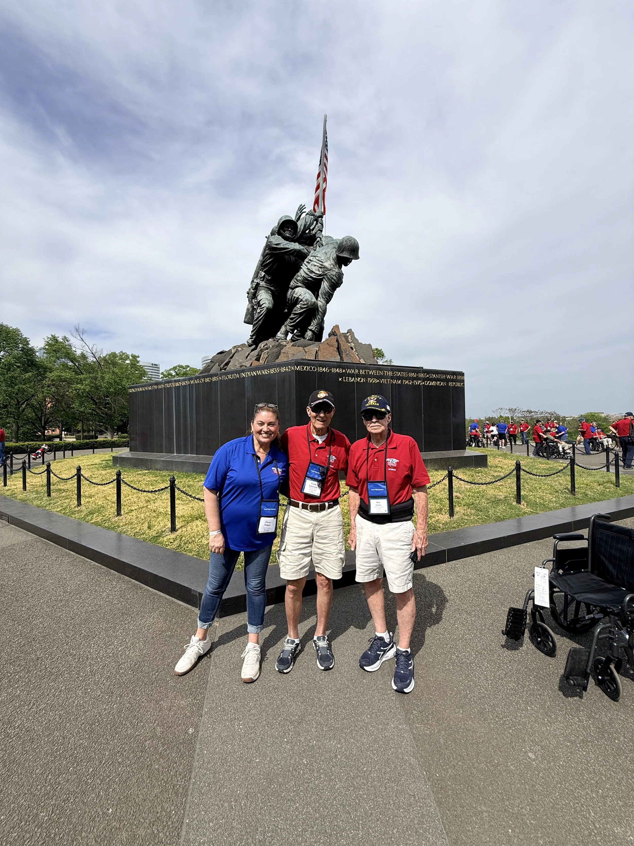 Three people, two men and a woman, standing in front of the Iwo Jima Memorial, smiling at the camera. The memorial features soldiers raising the American flag, with a group of people gathered around and others in the background.