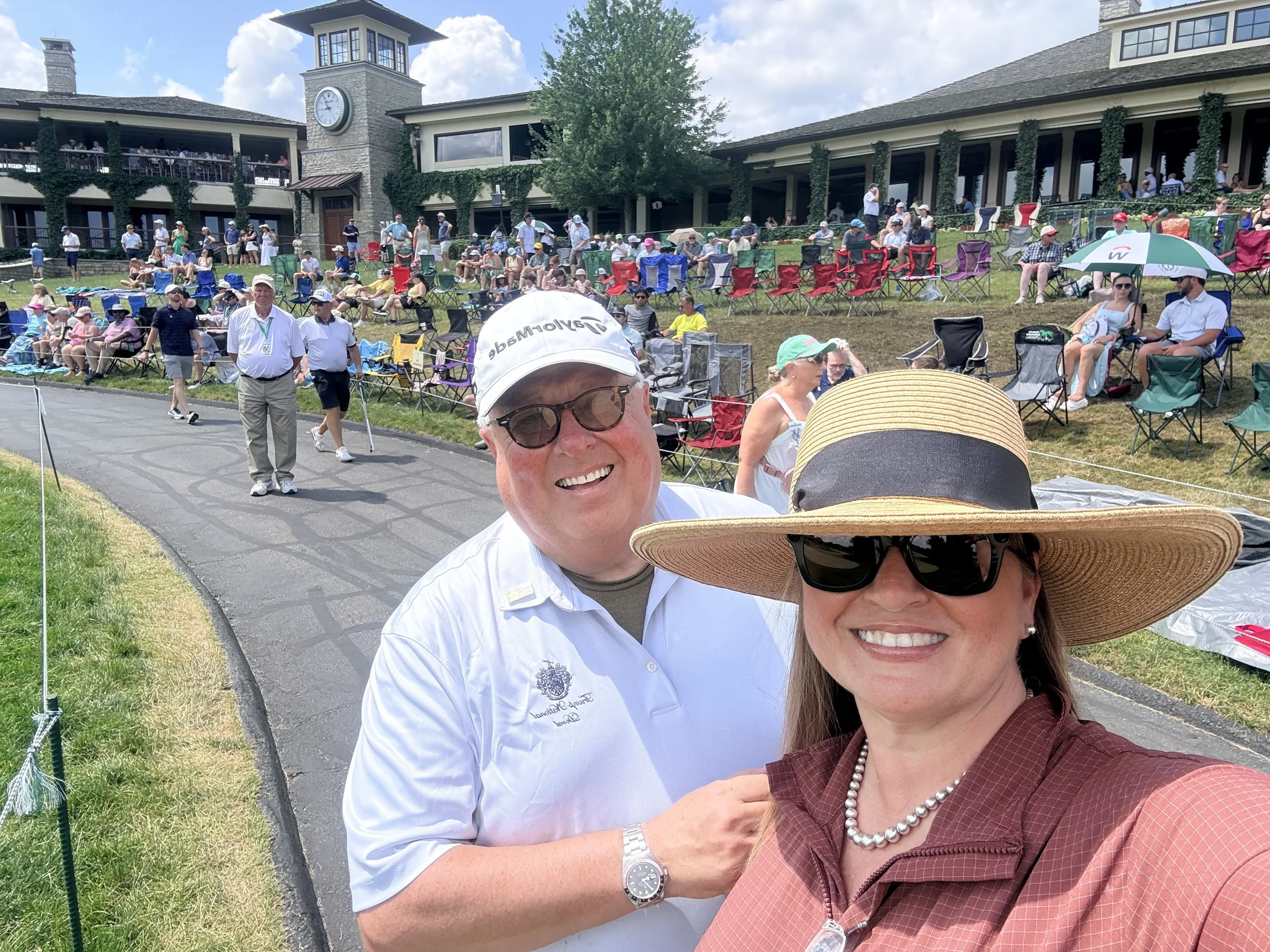 People sitting on chairs and standing on a grassy outdoor area of a golf course clubhouse, with some taking photos, wearing hats and casual summer clothing, on a sunny day with blue sky and clouds.