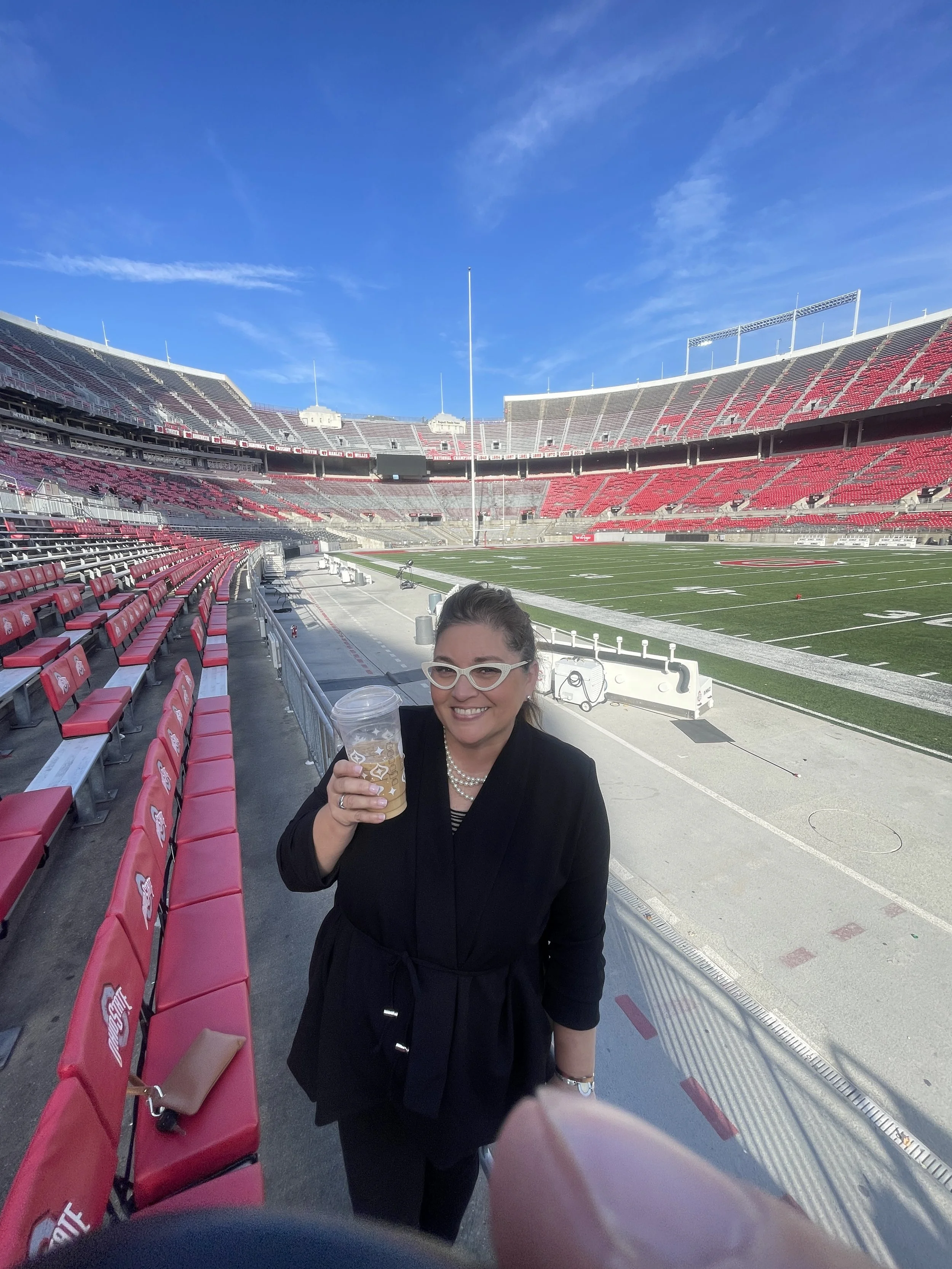 A woman in a black coat and glasses holding a cup of beer standing inside a large football stadium.