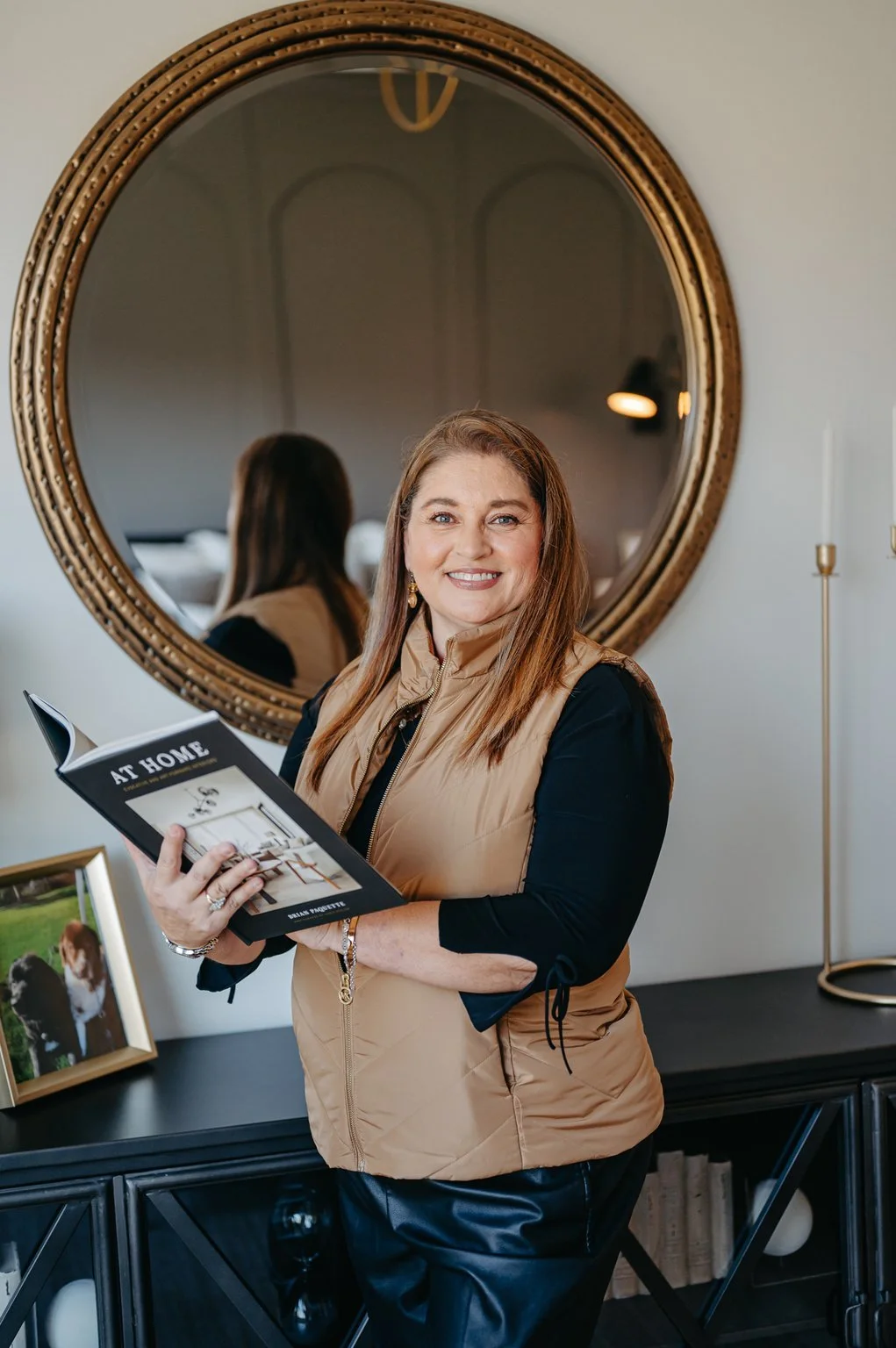 A smiling woman with shoulder-length brown hair holding a magazine titled "At Home," standing in front of a large round mirror with a gold frame. She is wearing a tan vest over a black shirt and is standing in a modern, well-decorated room.