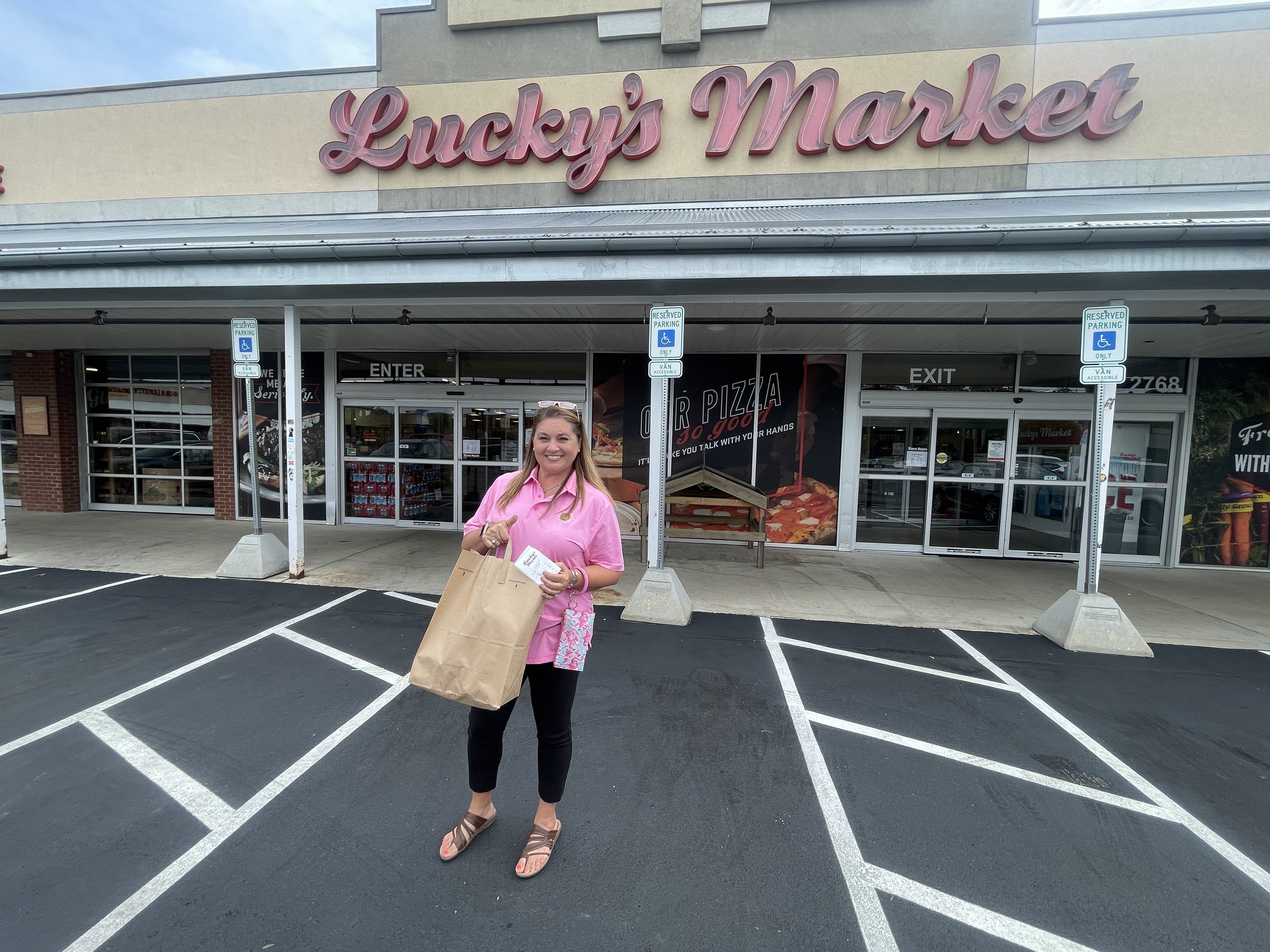 Woman standing in a parking lot holding a paper bag and smiling, in front of Lucky's Market store entrance with reserved parking signs for handicapped, and a woman wearing a pink shirt, black pants, and sandals.