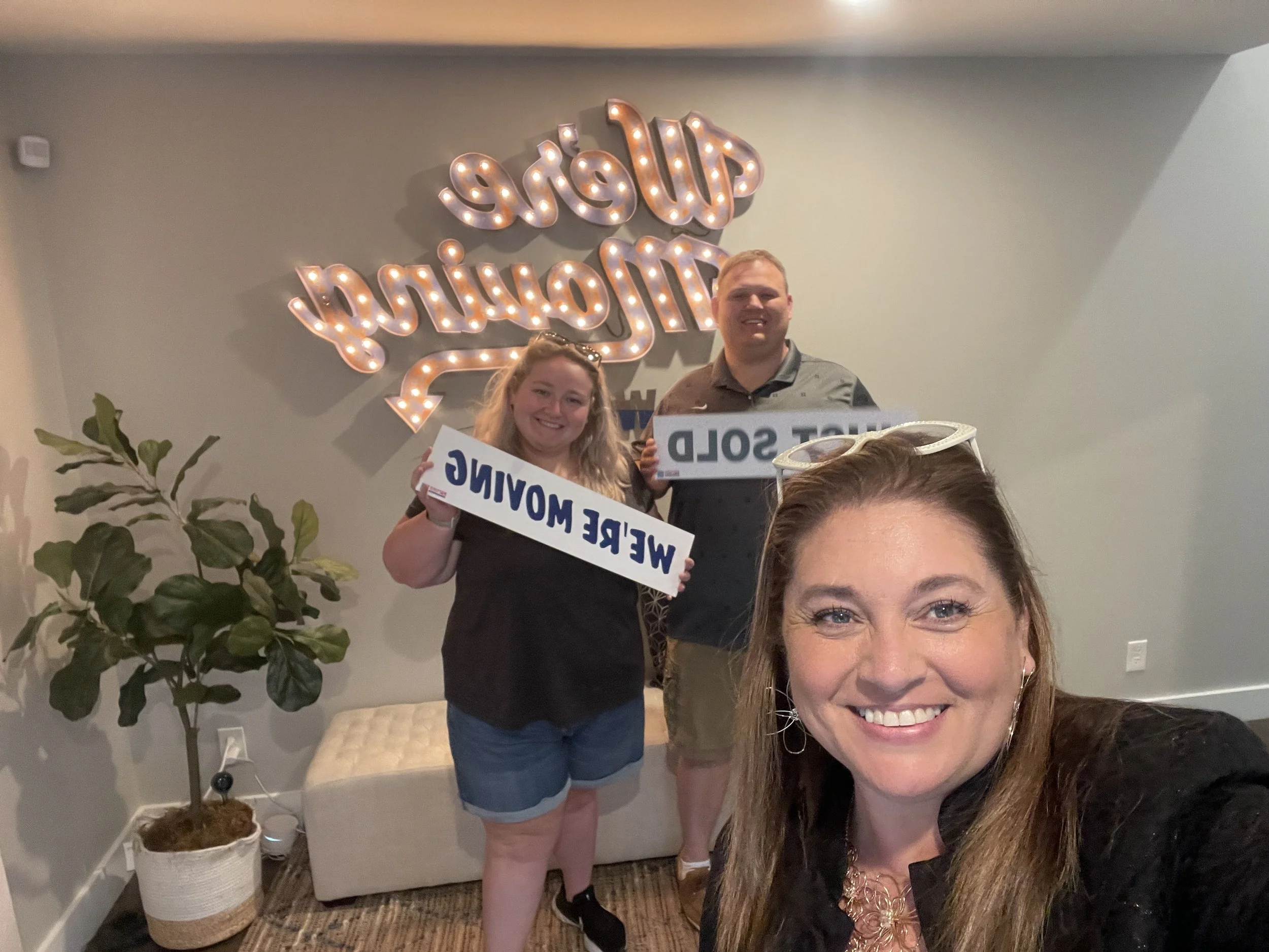 Four people posing for a photo at an indoor gathering. Behind them is a decorative sign with lights that reads 'Weld' and 'Dream'. The woman in the front is smiling, and the others are holding signs. There's a potted plant nearby, and the setting app