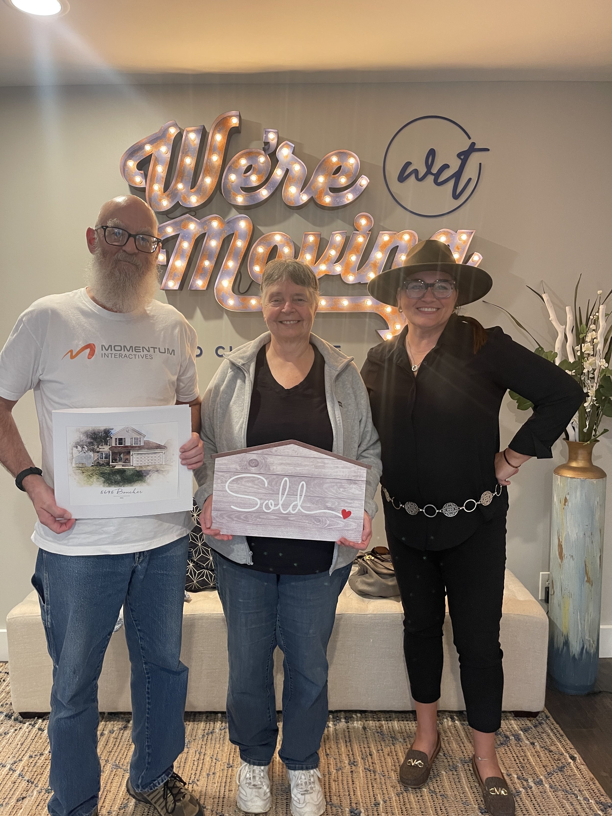 Three adults standing indoors in front of a wall with illuminated sign that reads 'Were Moving'. The person on the left is a man with glasses and a beard, wearing a white T-shirt and blue jeans. The middle person is a woman with short gray hair, wear