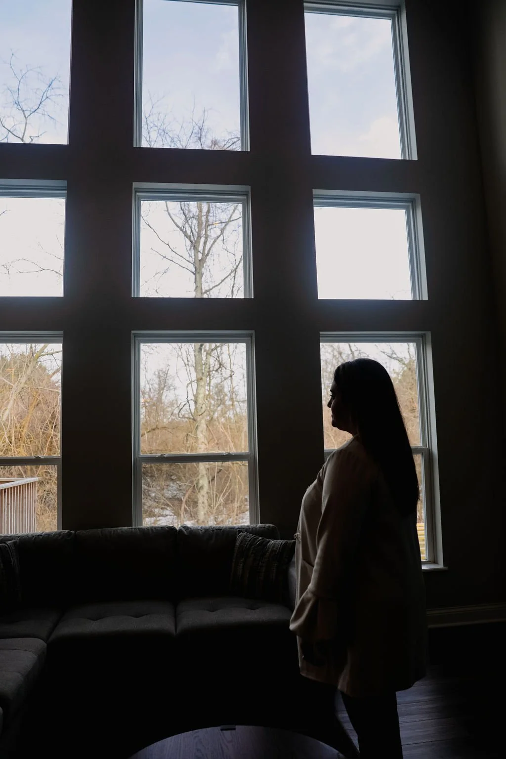 A woman stands in profile inside a room, looking out a large multi-pane window with a view of leafless trees outside during daytime.