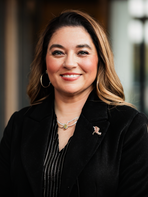 A smiling woman with wavy brown hair wearing a black blazer, layered necklaces, hoop earrings, and a small flag pin, standing indoors with blurred windows in the background.