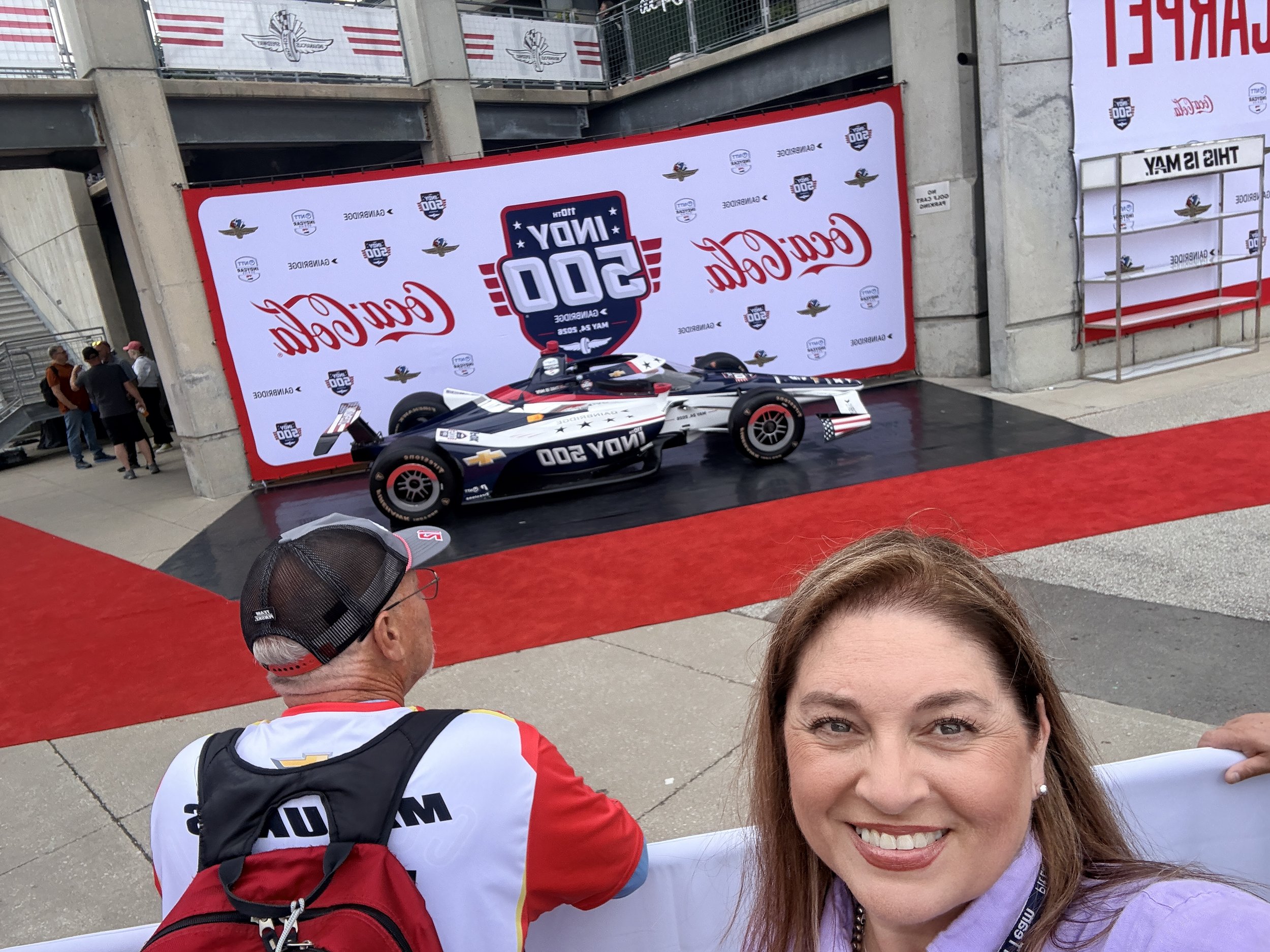 Smiling woman taking a selfie at a racing event, with race car and event banners in the background.