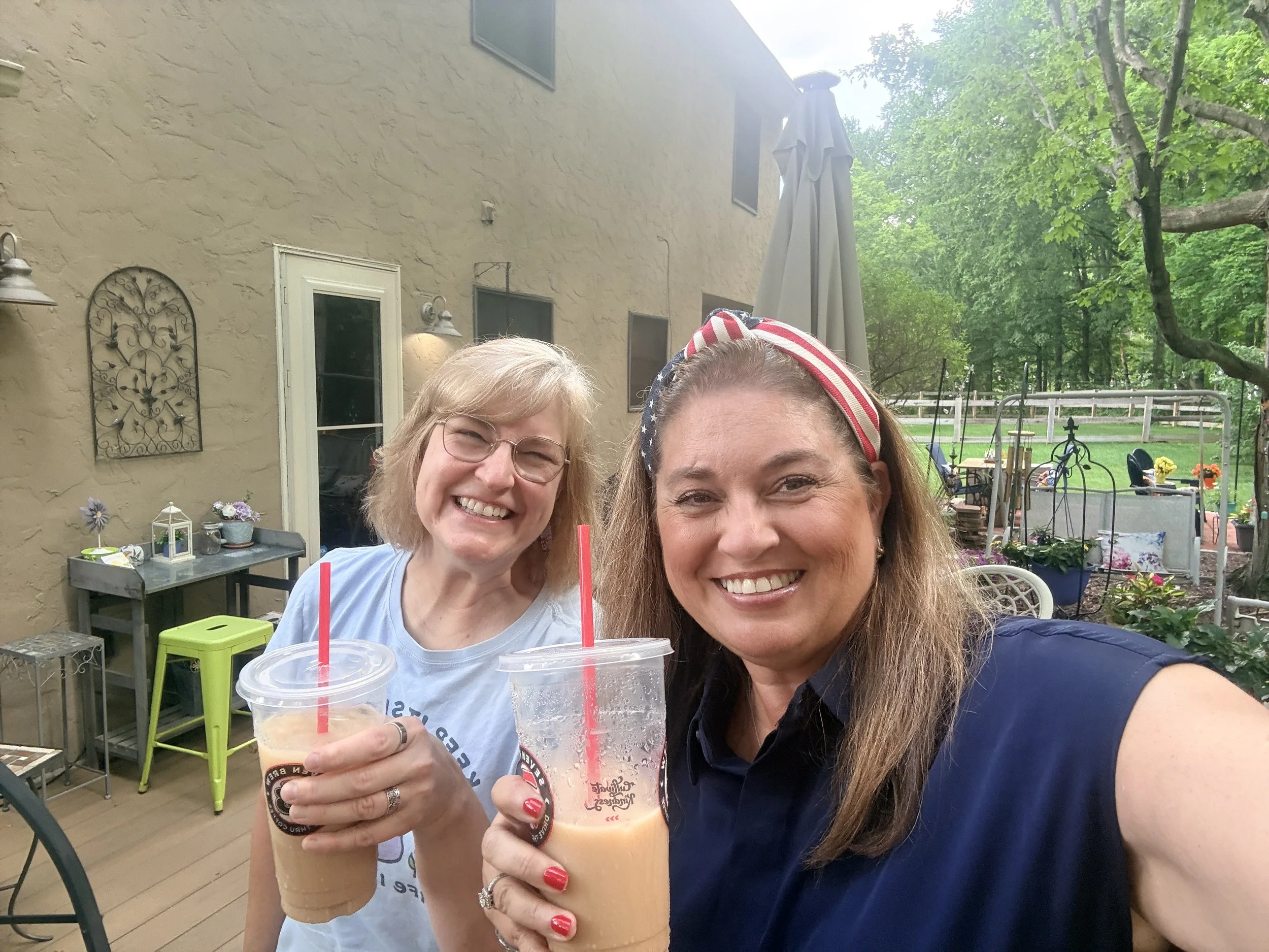 Two women smiling and holding iced coffee drinks with red straws in an outdoor patio area, with green trees, patio furniture, and a beige brick house in the background.