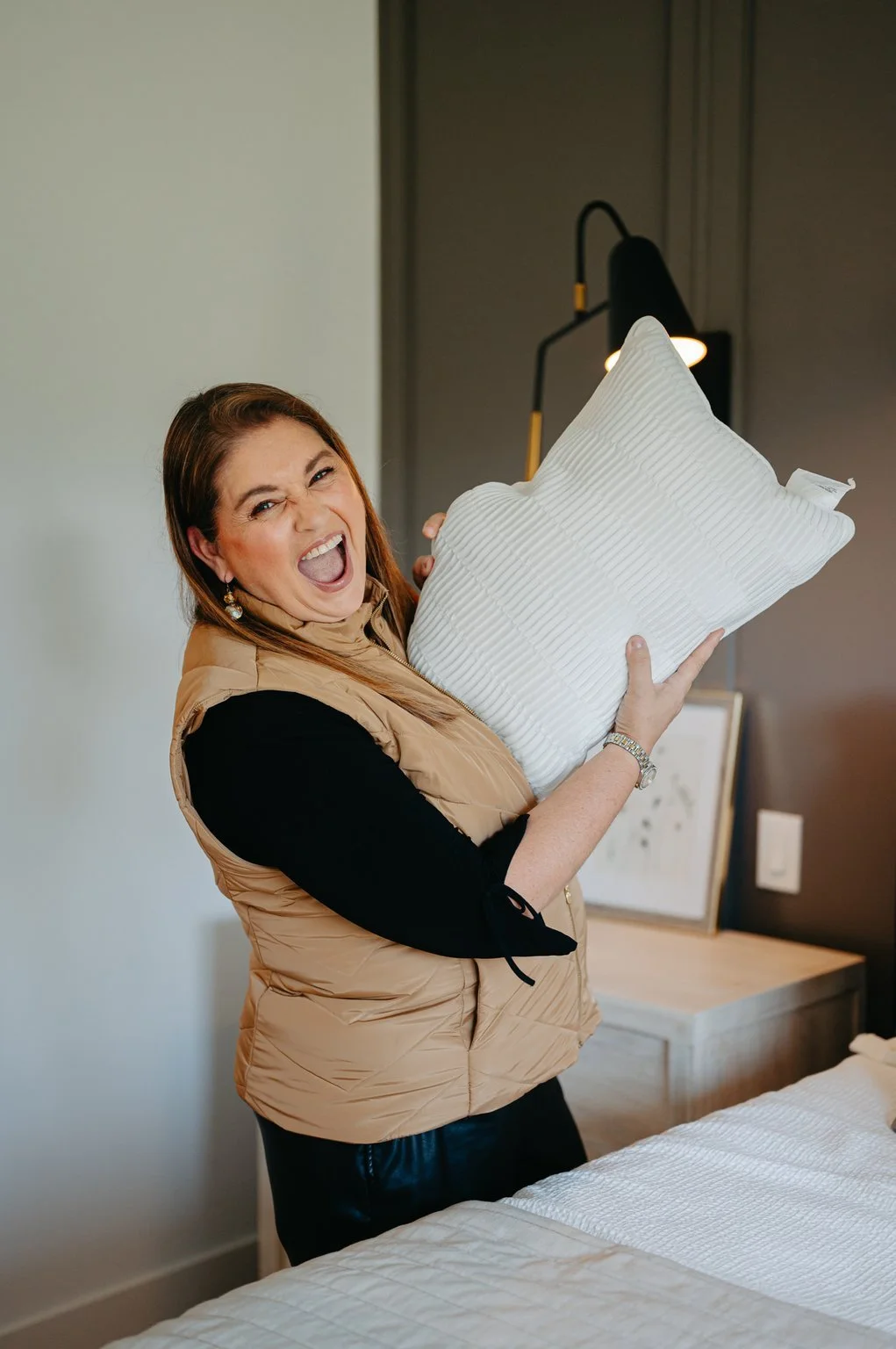 A woman holding a white pillow and smiling happily in a bedroom.