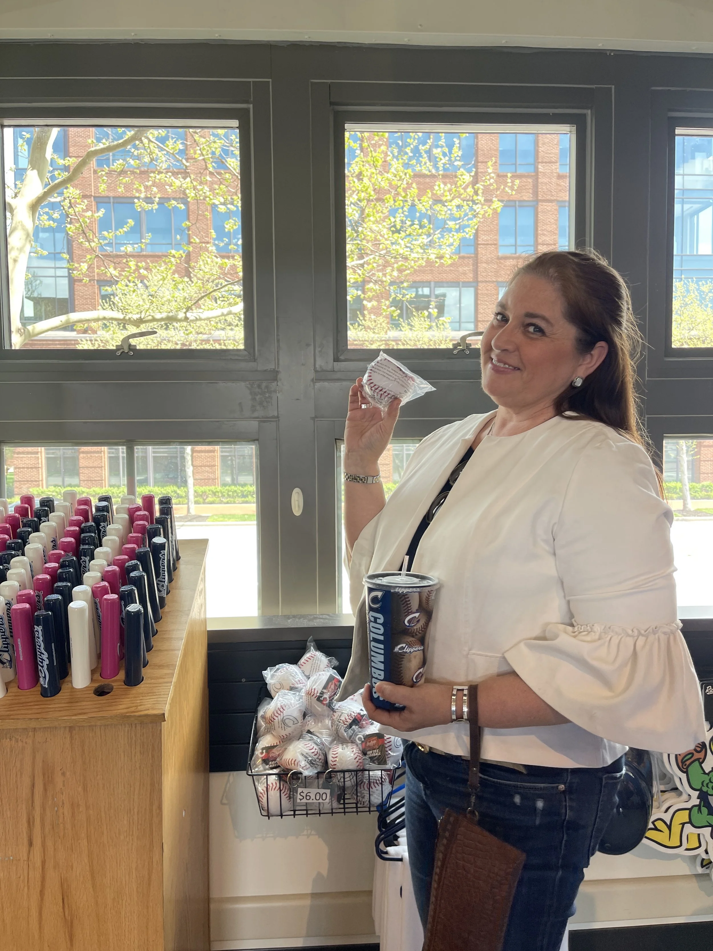 A woman in a white jacket holding a baseball ice cream bar and a cup of soda, standing near a display of pink, black, and white markers and a basket of baseballs with a $6.00 price tag, inside a building with large windows and a cityscape background.